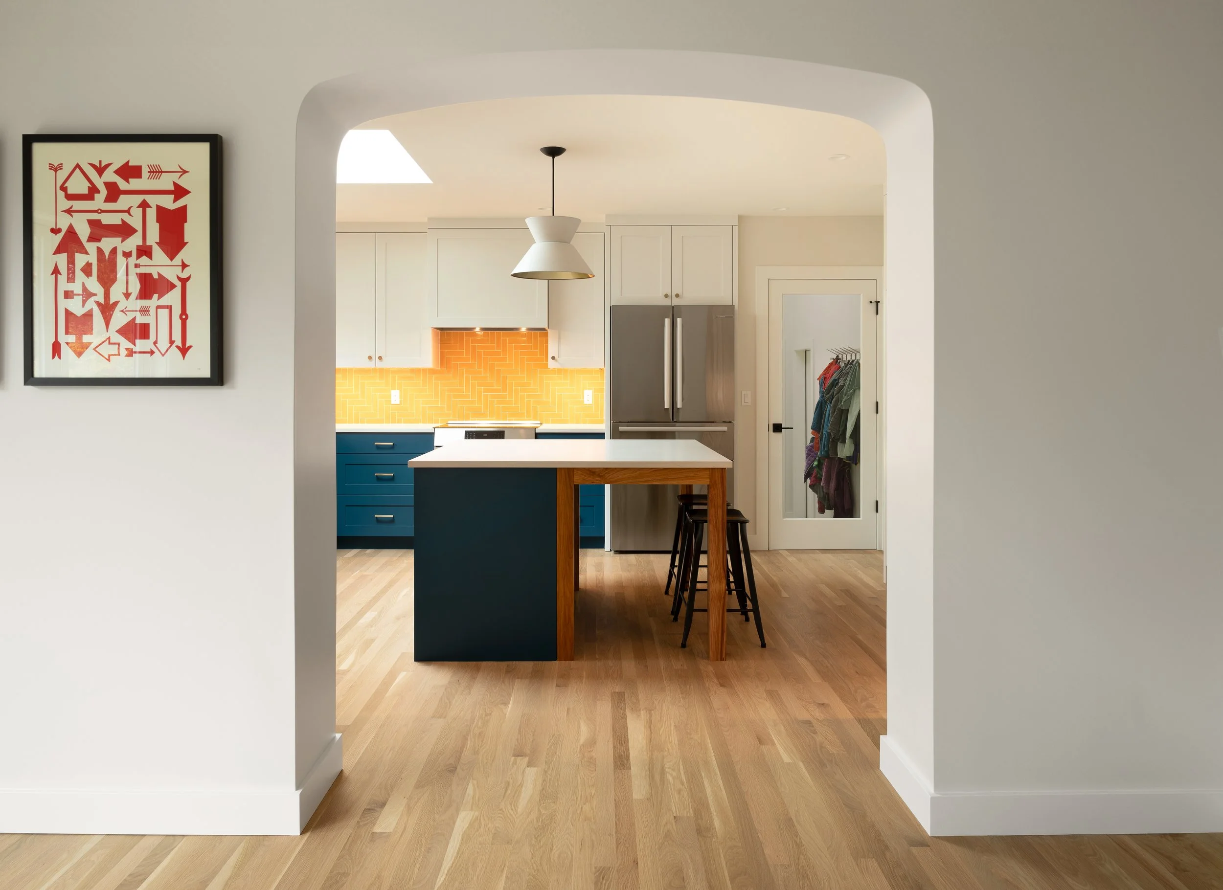 Remodeled kitchen through an archway, featuring white cabinets, a stainless steel refrigerator, a yellow backsplash, a suspended white pendant light, a kitchen island with bar stools, with a wall to the right displaying a coat hook and clothing.