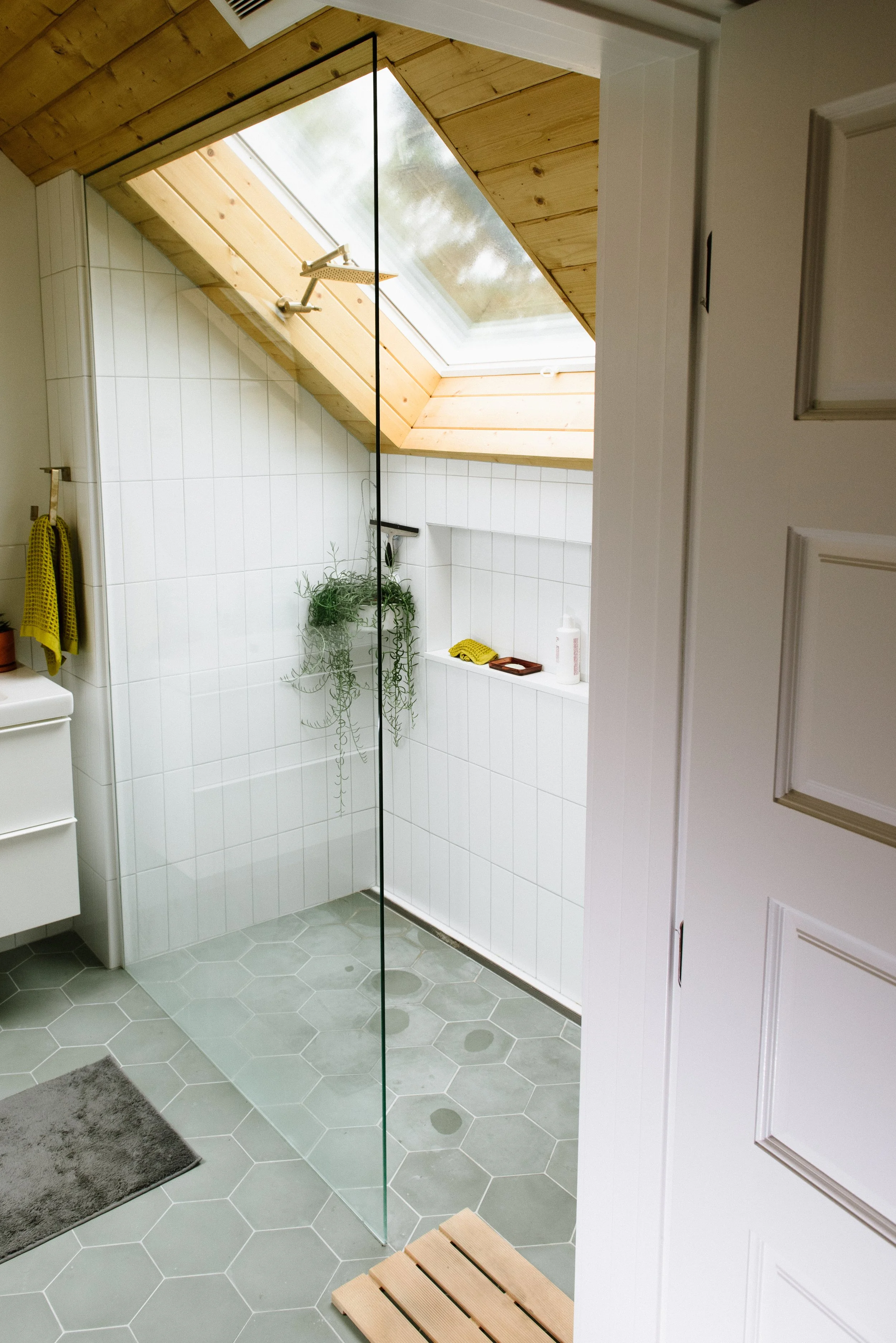 A bathroom with a wooden ceiling and a skylight window, a glass shower door, white tiled walls, and a gray hexagon tile floor. There is a small wooden bench outside the shower, a gray bath mat, a towel, and some toiletries.