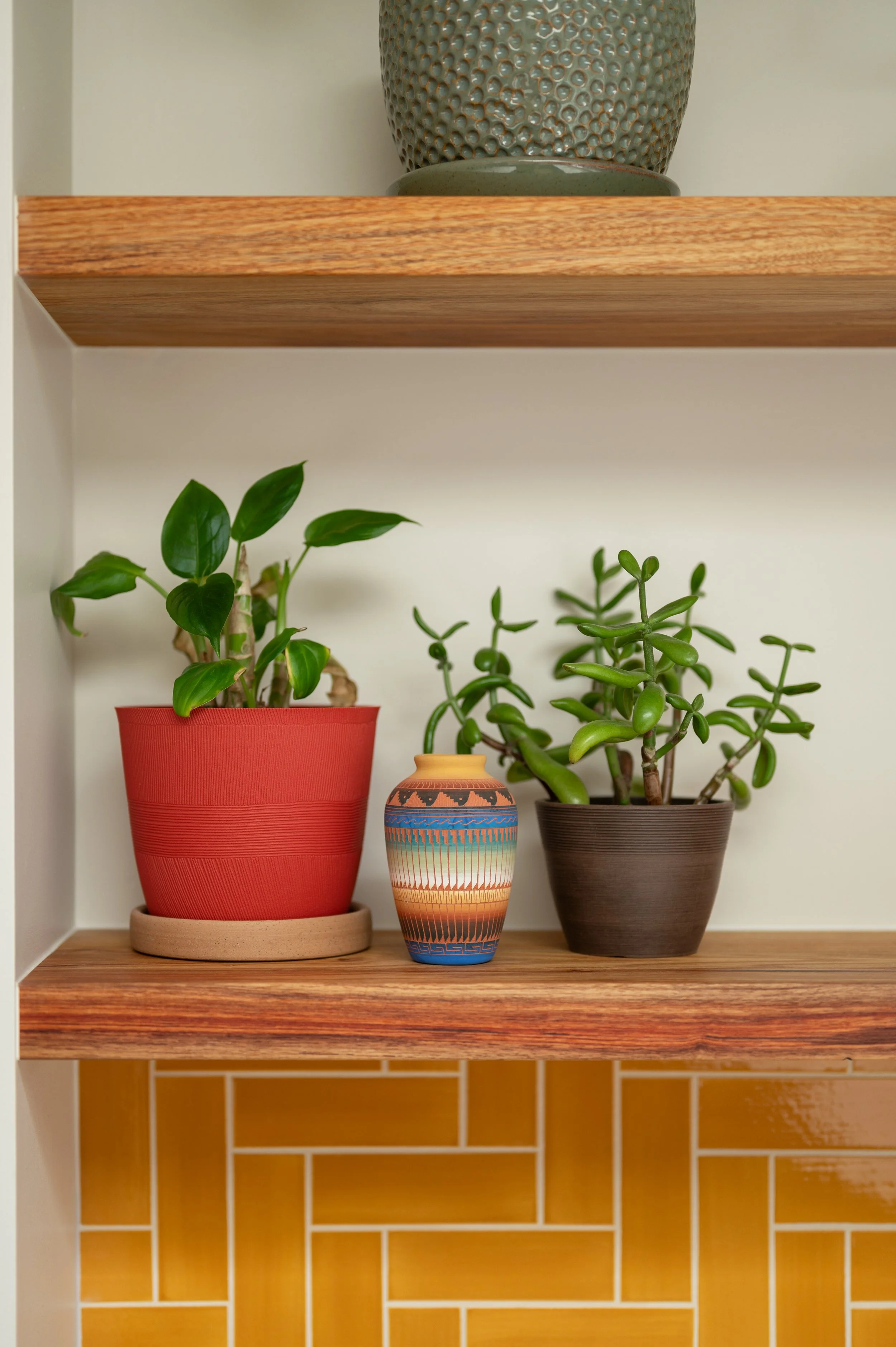Detail of three potted plants on a wooden shelf with a decorative vase in the middle. The shelf has a second level above and a yellow brick background below.