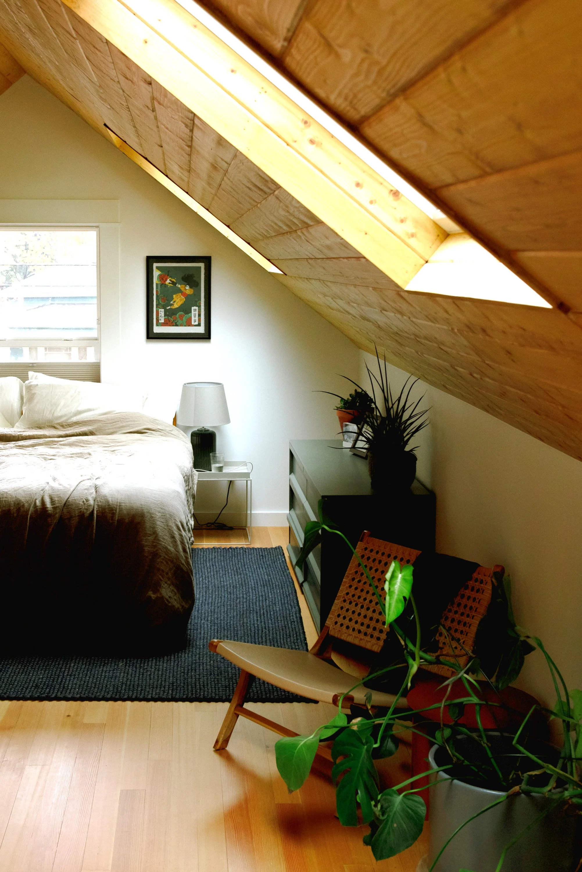 Two skylights and a window above the bed, with small nightstand holding a lamp, and dresser in this renovated attic bedroom.