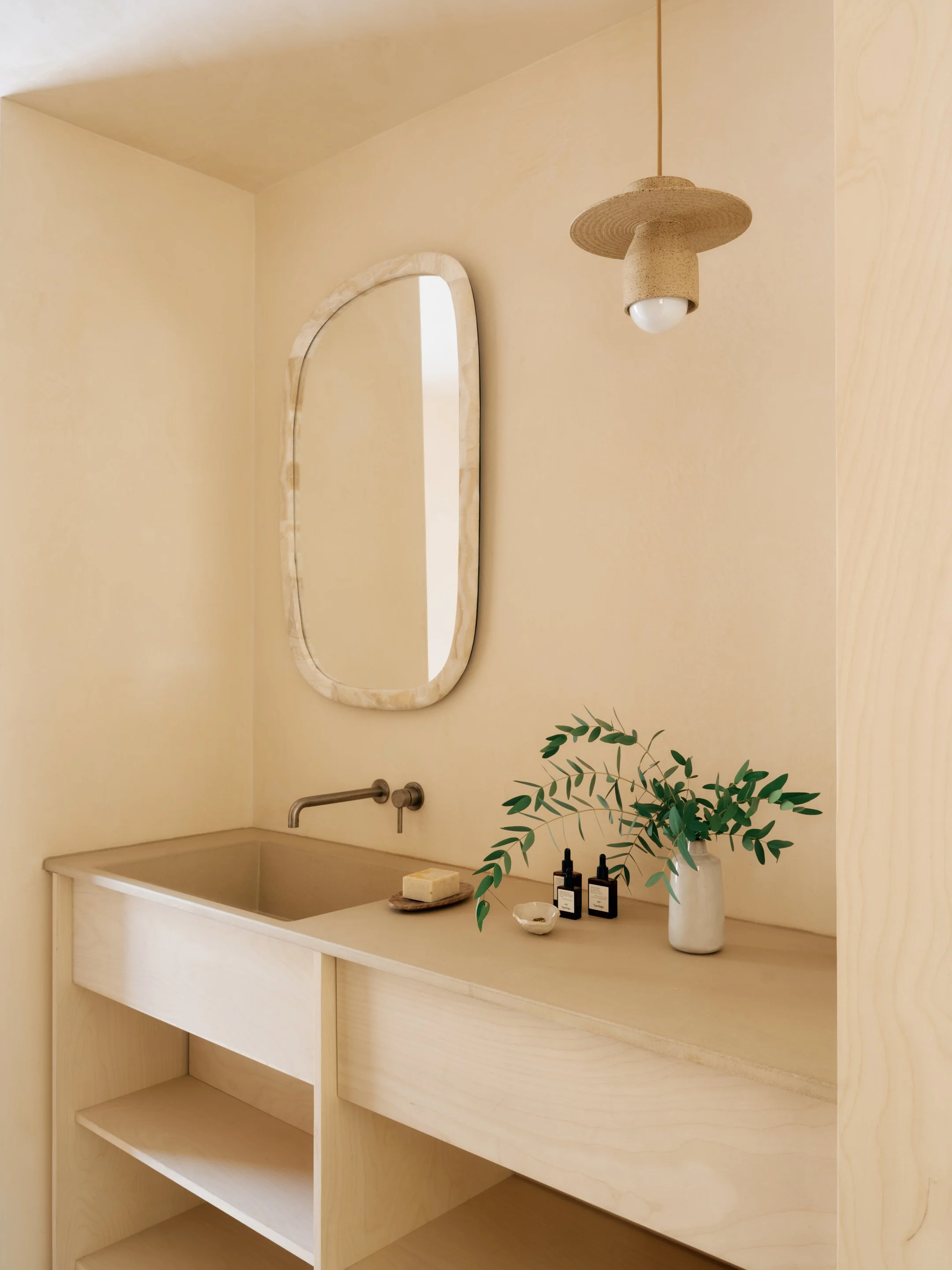 A minimalist bathroom vanity with a beige countertop, an integrated sink, a modern wall-mounted faucet, decorative soap and lotion bottles, a white vase with greenery, a small dish, a soap bar, oval mirror with a marble frame, and a pendant light han