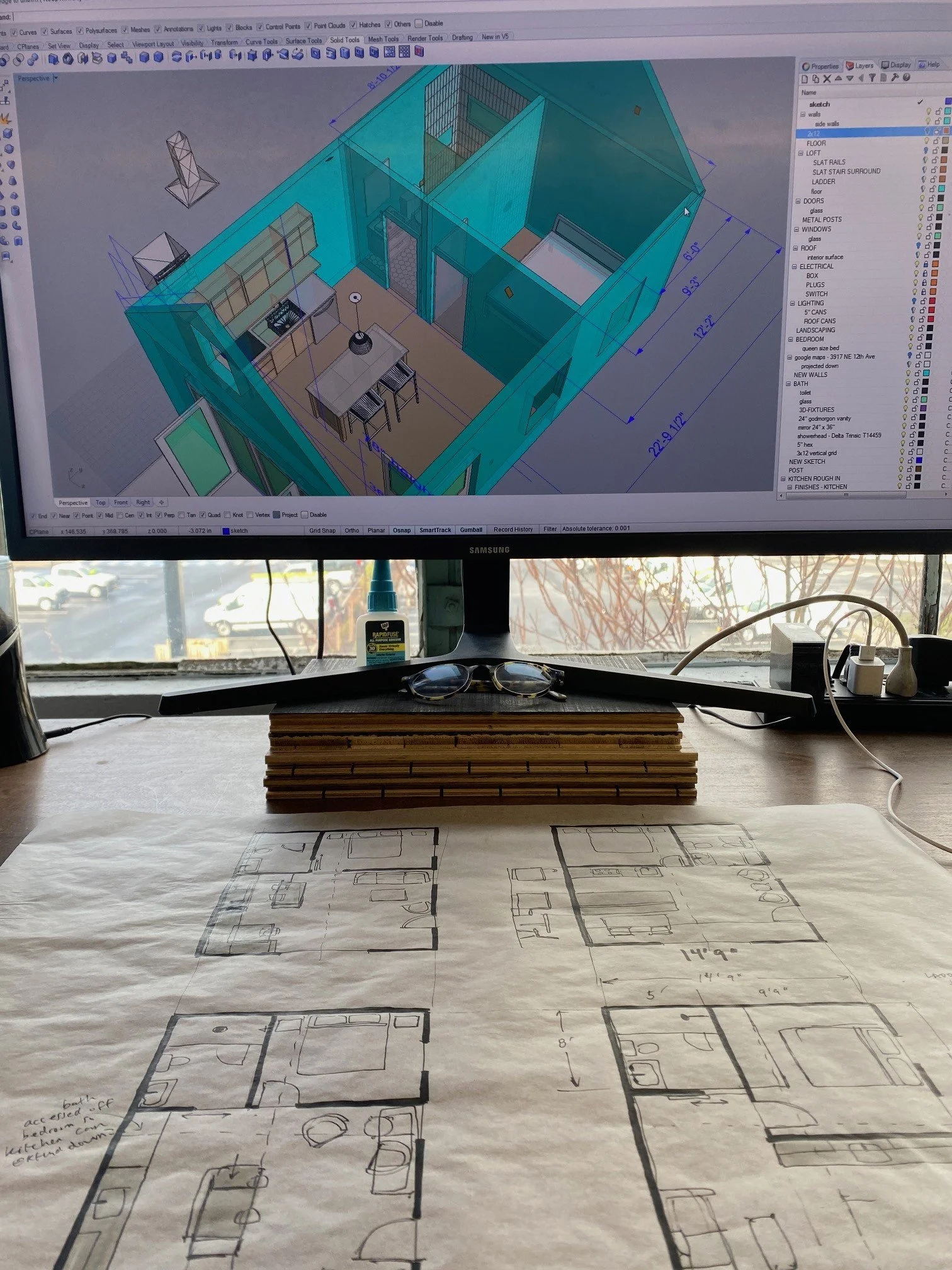 Computer screen displaying a 3D architectural design software with a house plan. The workspace includes blueprints, measurements, and various building components, with physical house plans and drawings on the desk below.