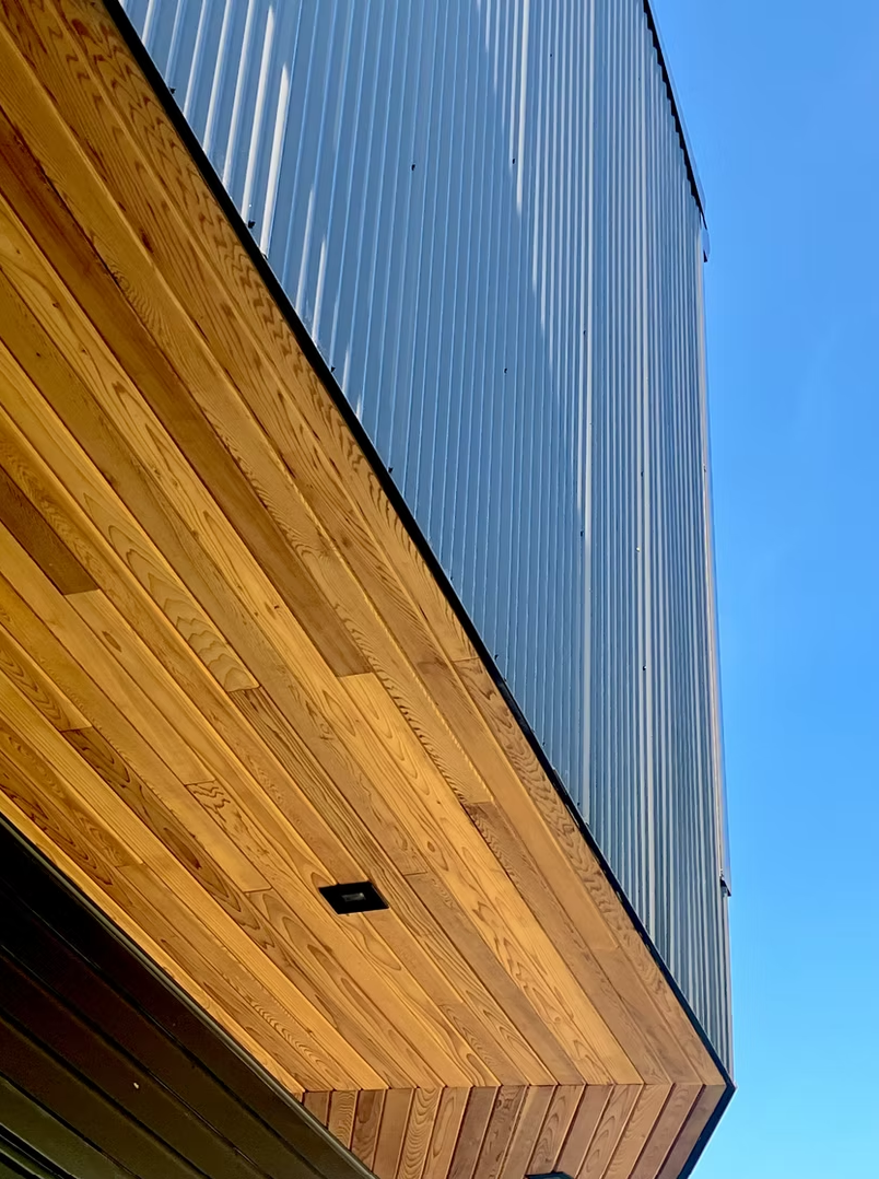 Upward view of a modern building with a wooden overhang and blue metal siding, against a clear blue sky.