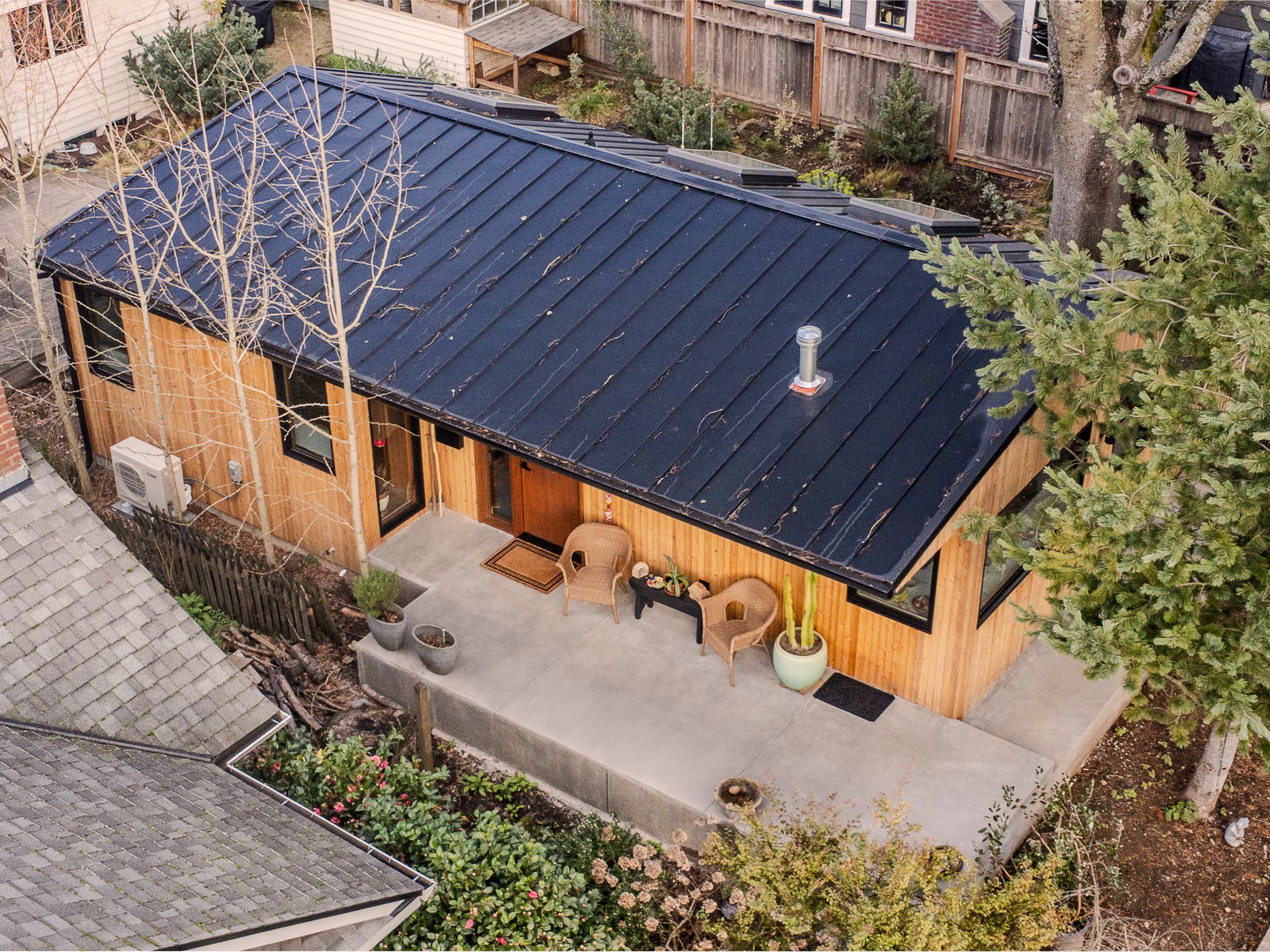 Aerial view of a backyard with a black metal roof ADU, surrounded by trees, plants, and a wooden fence, featuring a backyard patio area with two chairs, potted plants, and a small outdoor rug.