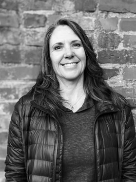 A woman with long hair smiling, wearing a dark jacket, standing in front of a brick wall.