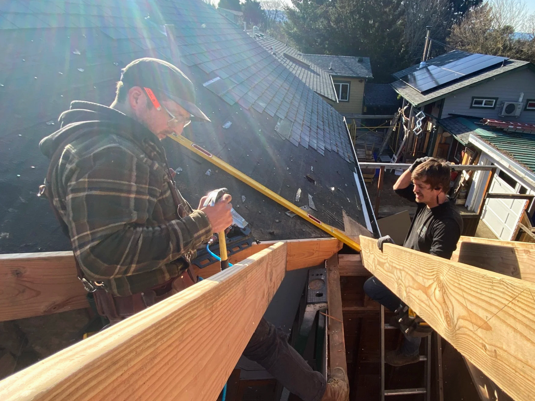 Two men working on a wooden framing project on the roof of a house during daylight. One man is using a level, and the other is smiling, both wearing gloves.