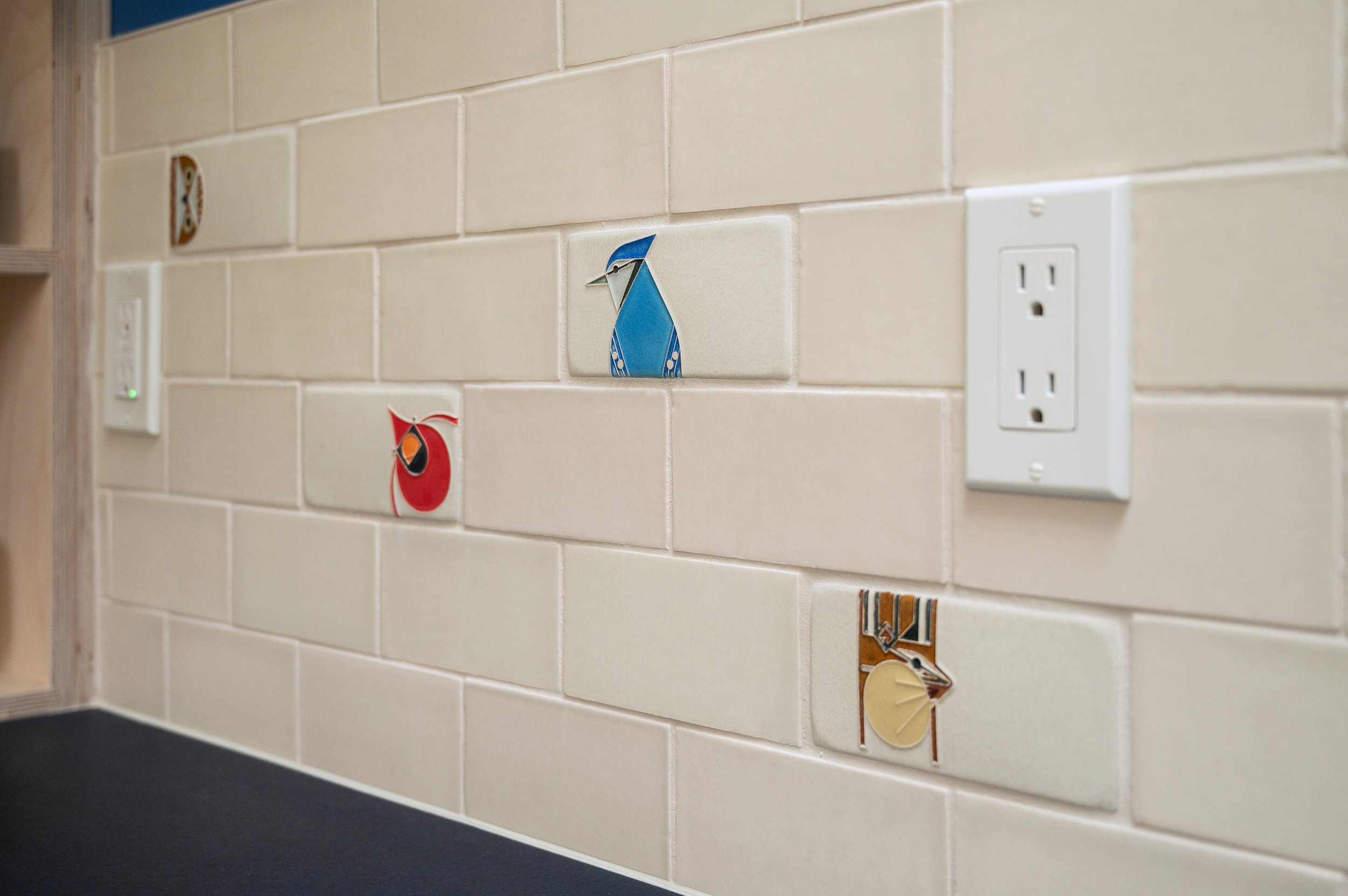ADU kitchen with beige tiled wall with various decorative tile pieces featuring geometric and animal designs, next to a white electrical outlet.