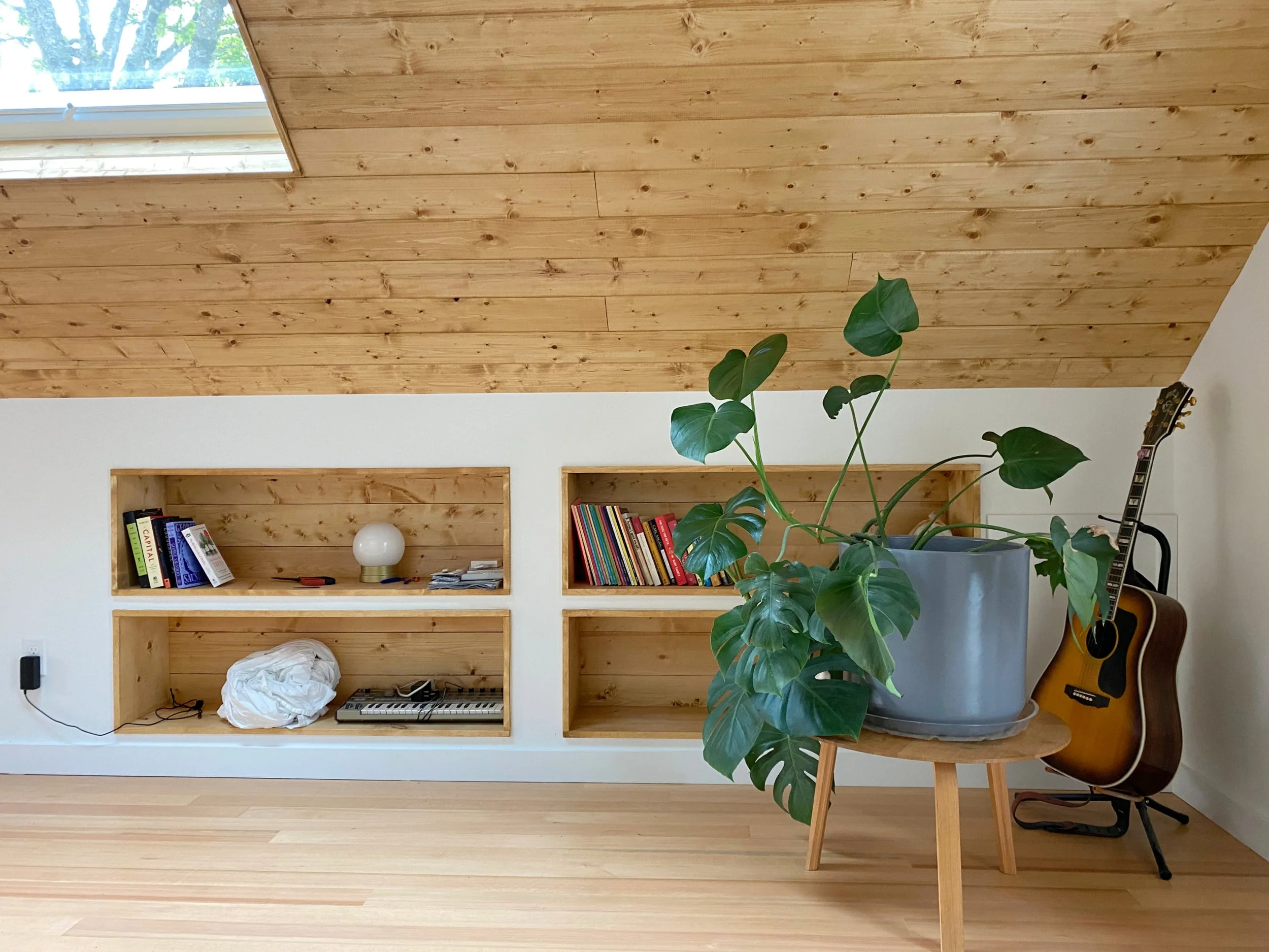 A wooden ceiling with a skylight window, built-in shelves with books and decorative items, a large green potted plant, an acoustic guitar, and a small wooden stool on a light wood floor.