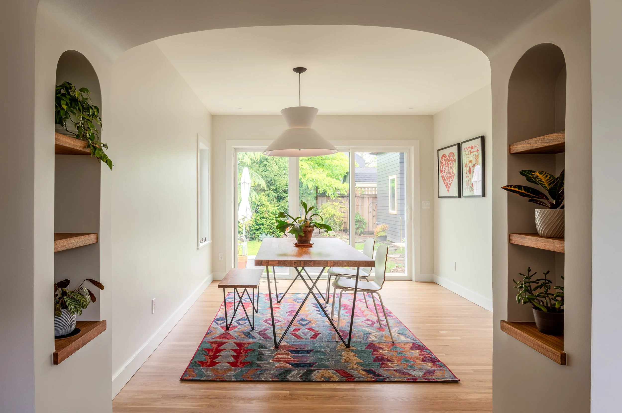 A bright remodeled dining area with a wooden table and metal chairs, a hanging white pendant light, a colorful geometric rug, and large sliding glass doors leading to a backyard with trees and a patio umbrella.