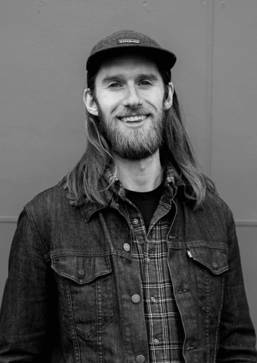 A man with long hair, a beard, and a baseball cap smiling, wearing a denim jacket over a plaid shirt, standing against a plain wall.