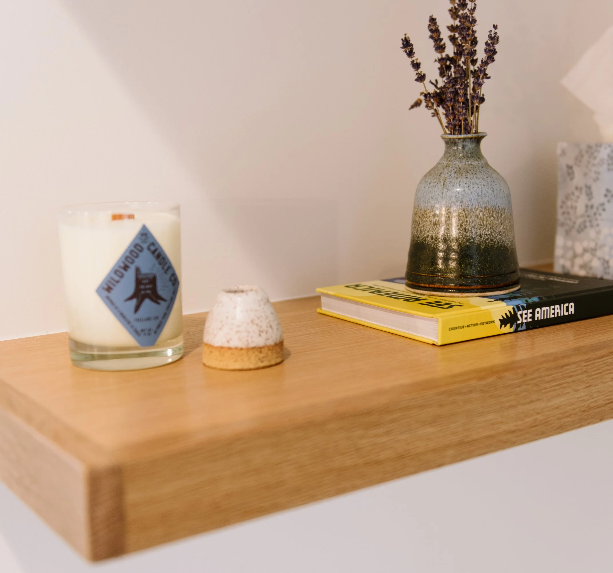 A wooden floating shelf with a candle, a small ceramic vase with dried lavender flowers, and a stack of two hardcover books.