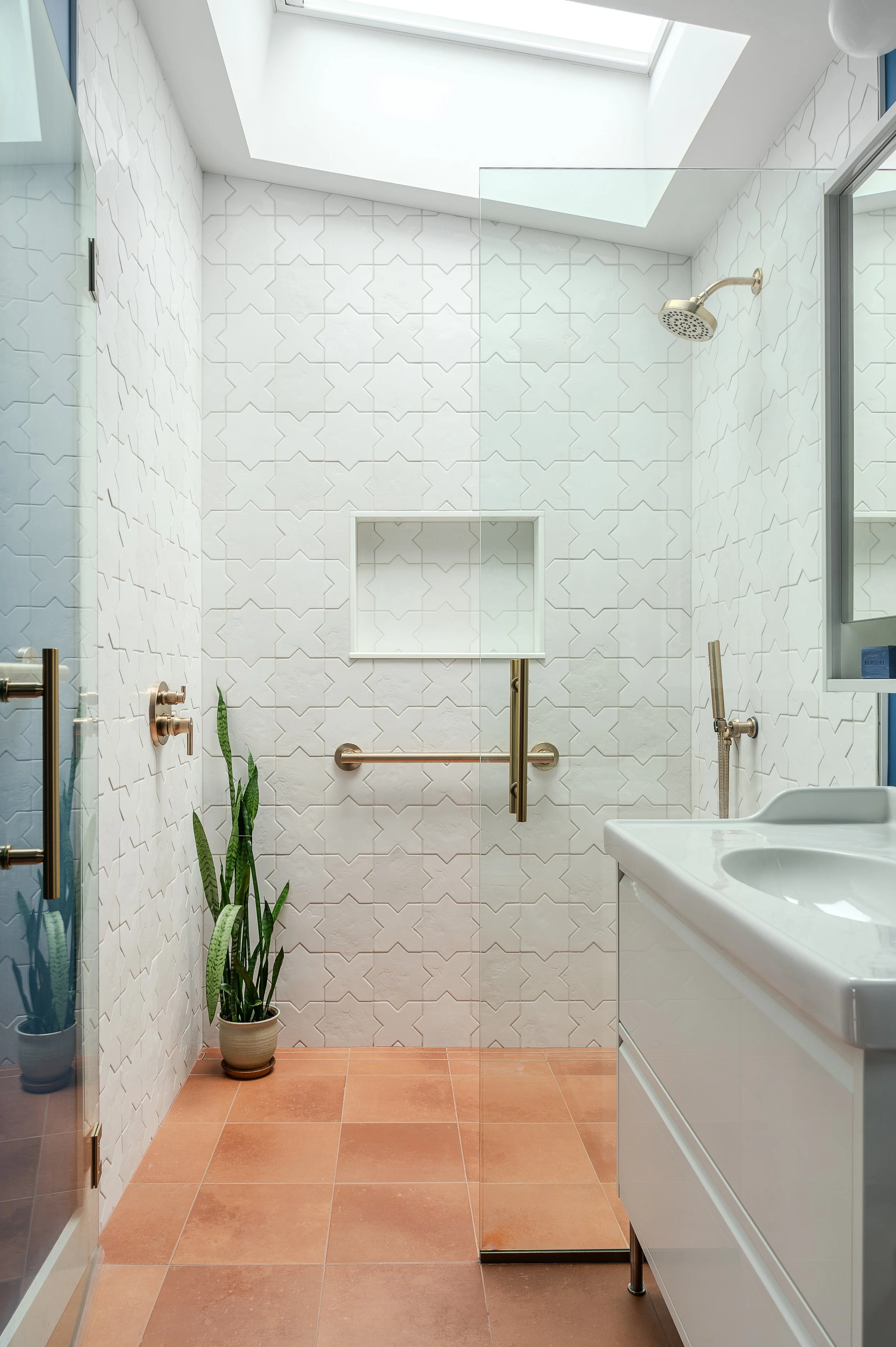Modern ADU bathroom with white patterned tiles, a sky skylight, a no-threshold shower with a glass door, a white vanity sink, a mirror, a tall green plant in a pot, and terracotta tile flooring.