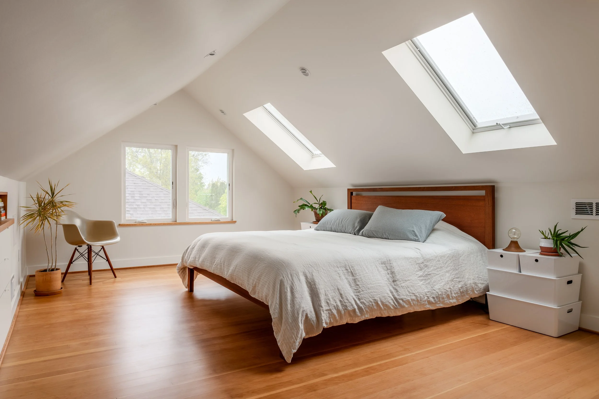 A remodeled attic bedroom with a white bed, two gray pillows, and wooden headboard. There are two skylights on the sloped ceiling, two windows with a view of trees, and hardwood floor.