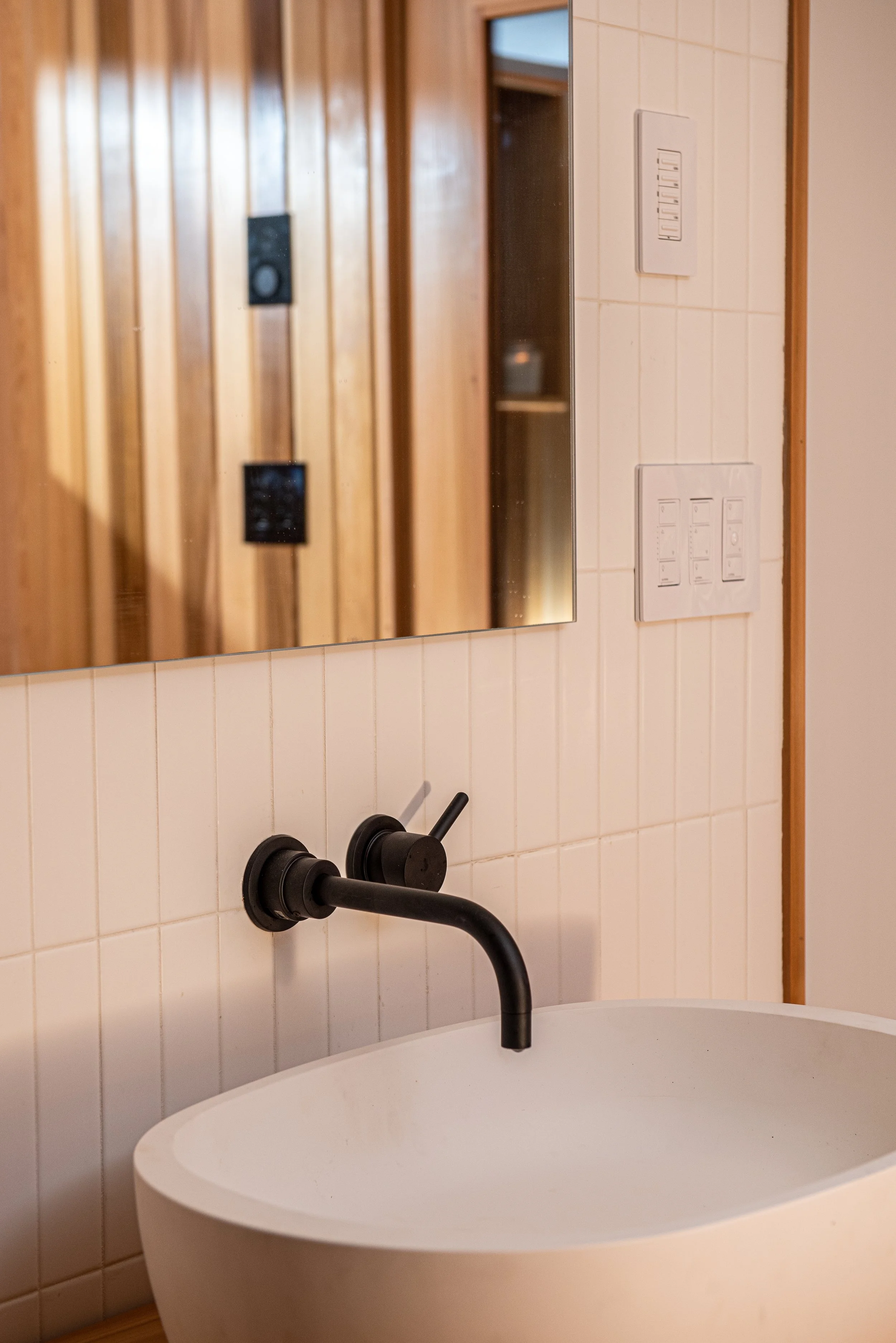 In this remodeled basement sauna, the white basin sink sits on a sturdy wooden shelf, with a dark faucet, and switch plates on the wall next to the frameless rectangular mirror above the sink. 