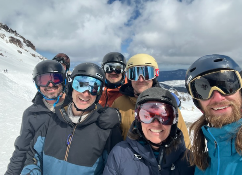 Group of six people in ski gear smiling on snowy mountain with cloudy sky