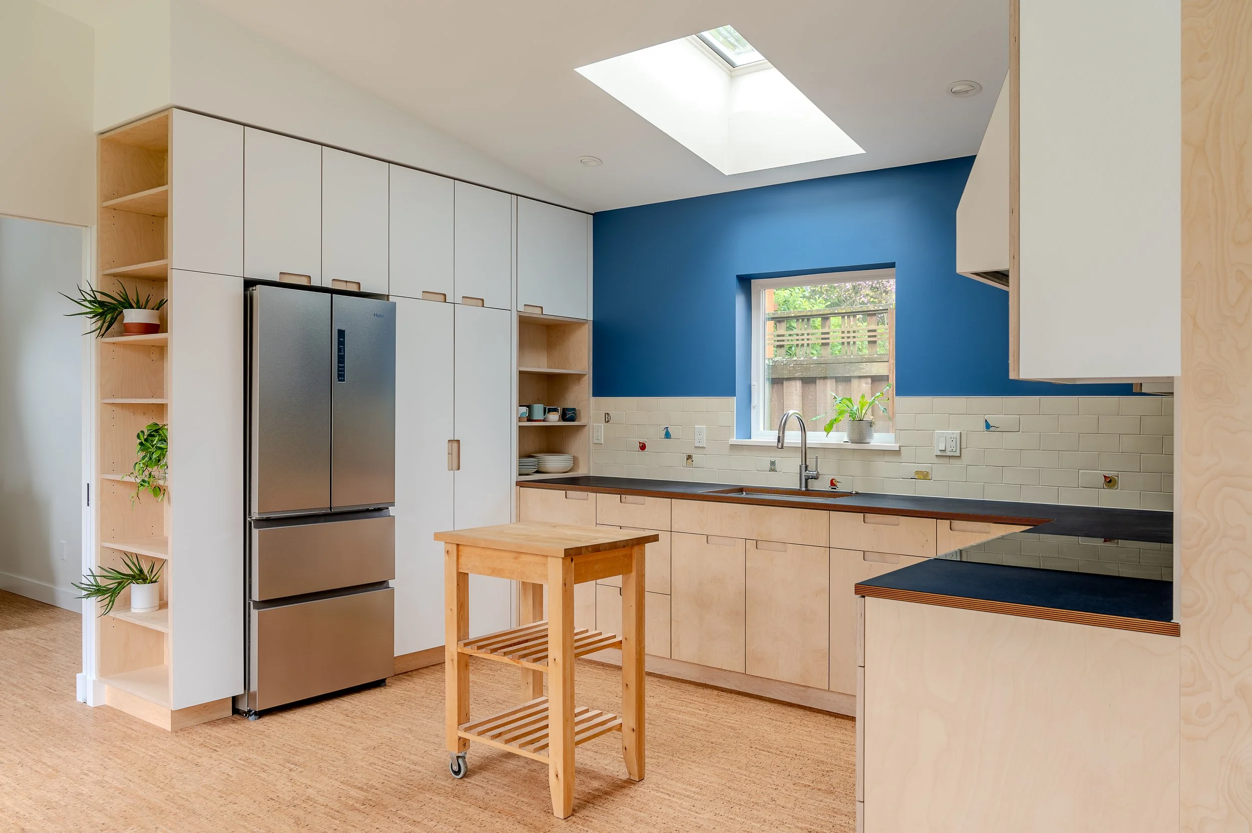 Modern ADU kitchen with white cabinets, a blue accent wall, a skylight, and a window with a view of a backyard.