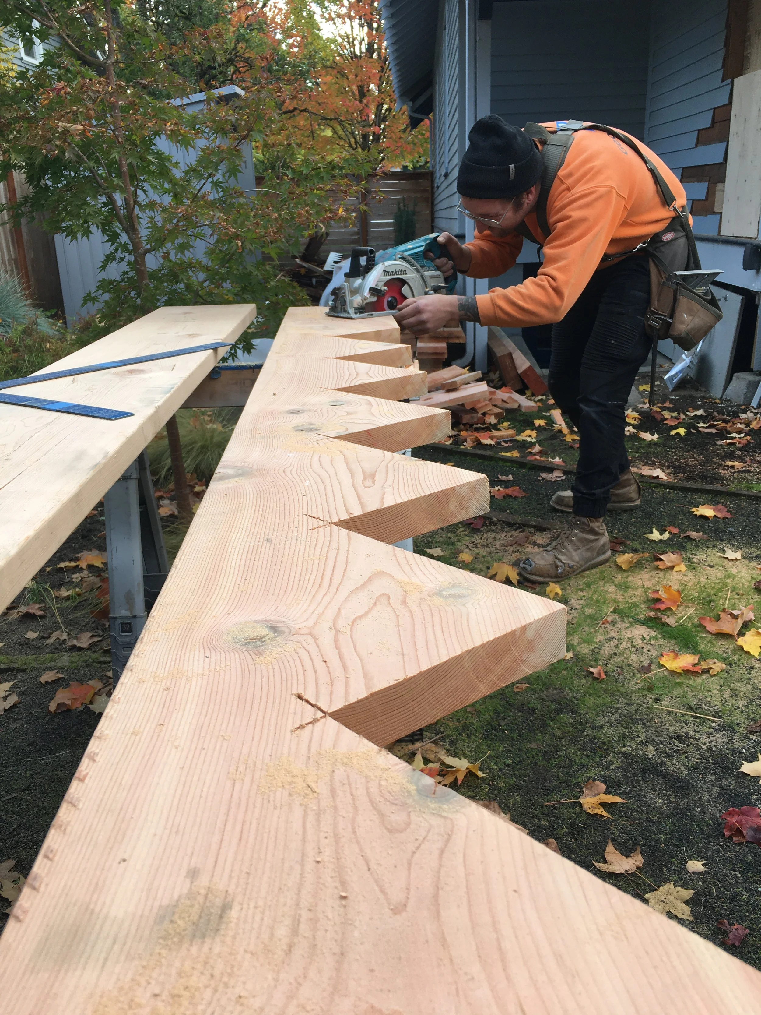 A construction worker in long-sleeved orange shirt using a circular saw to cut a piece of wood outside, with autumn leaves on the ground and trees in the background.