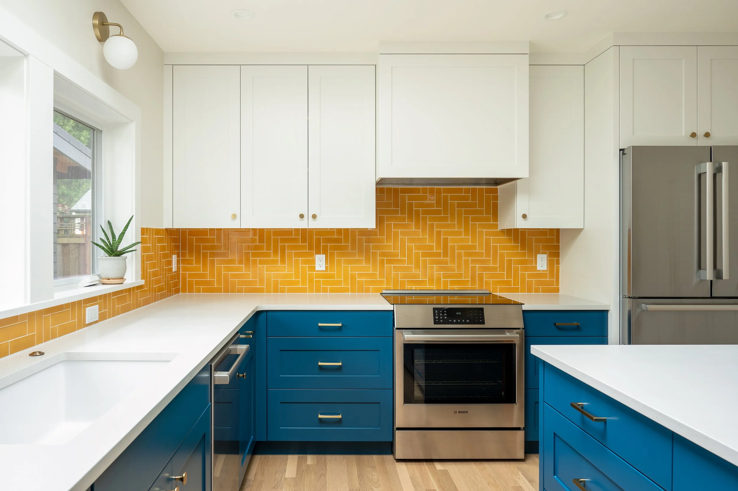 Modern ADU kitchen with white upper cabinets, blue lower cabinets, canary yellow tile backsplash in a herringbone pattern, stainless steel stove and refrigerator, window with a plant on the sill.