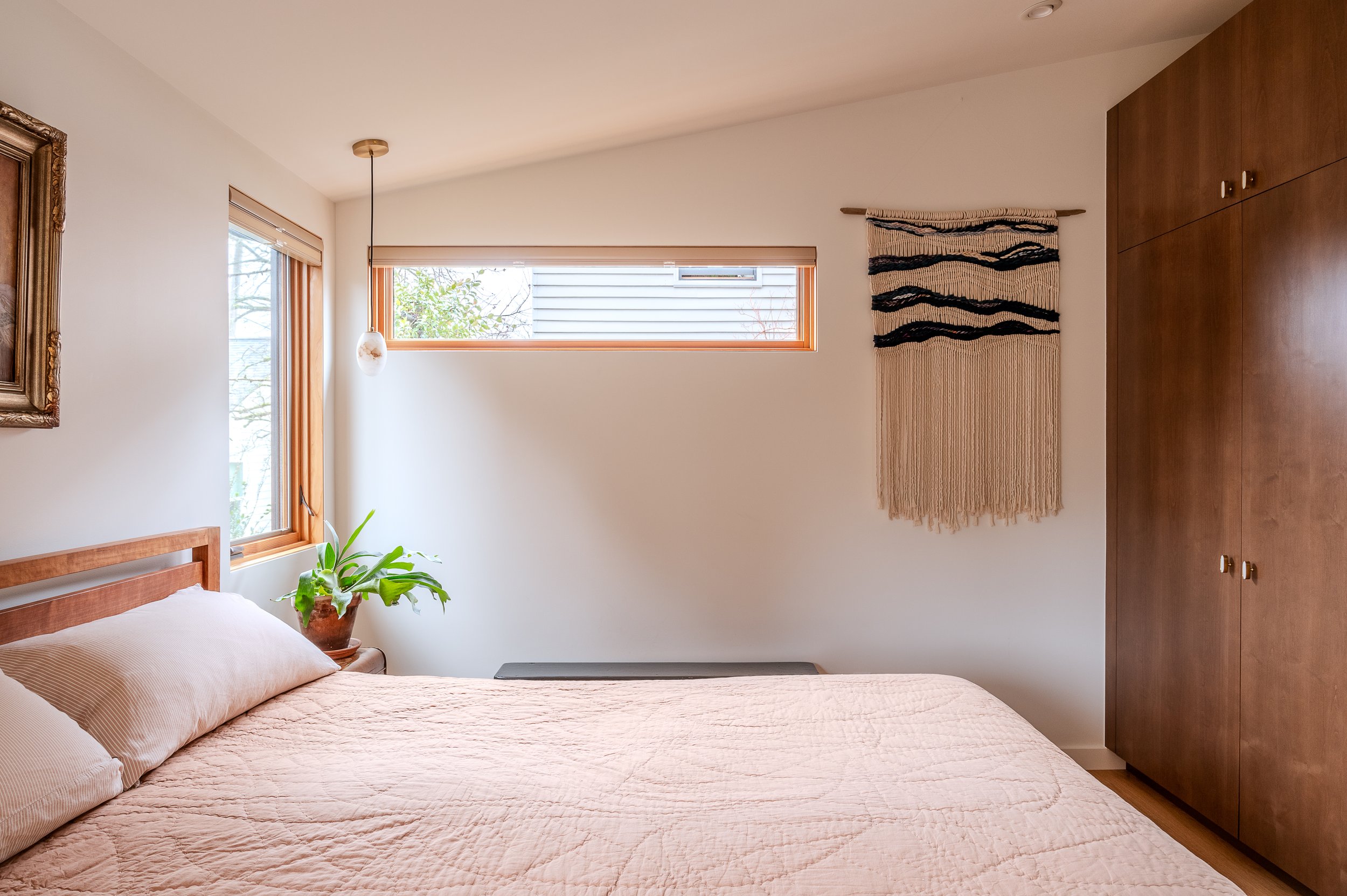 A cozy ADU bedroom with a bed, beige quilt, and matching pillows. There are two windows letting in natural light, one tall and one long and narrow. A potted plant on a side table, a wall hanging with black and beige design, a light pendant, and a woo