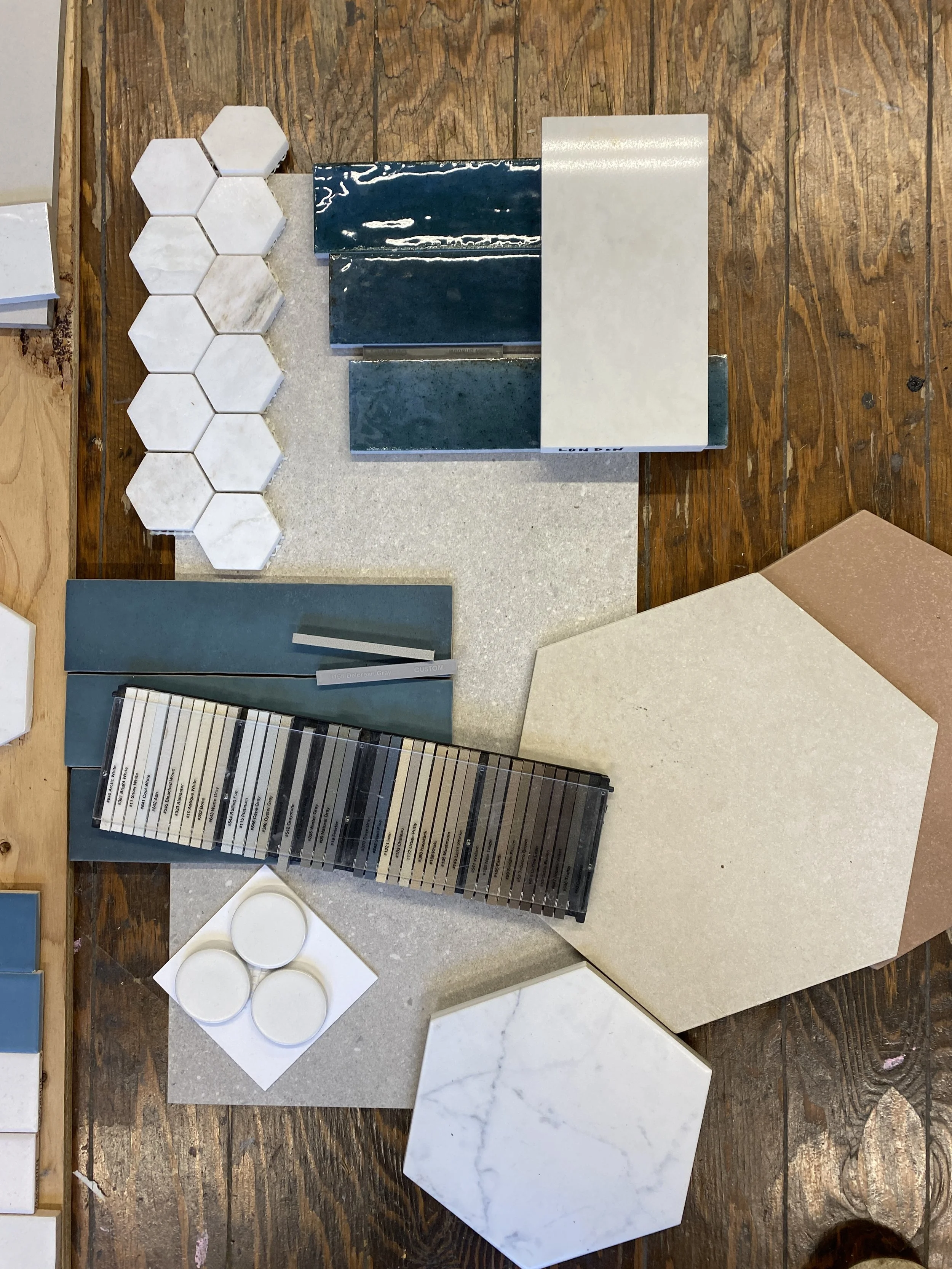 Samples of various tiles and materials on a wooden table, including hexagonal white tiles, blue glazed rectangular tiles, marble-like slabs, and color samples.