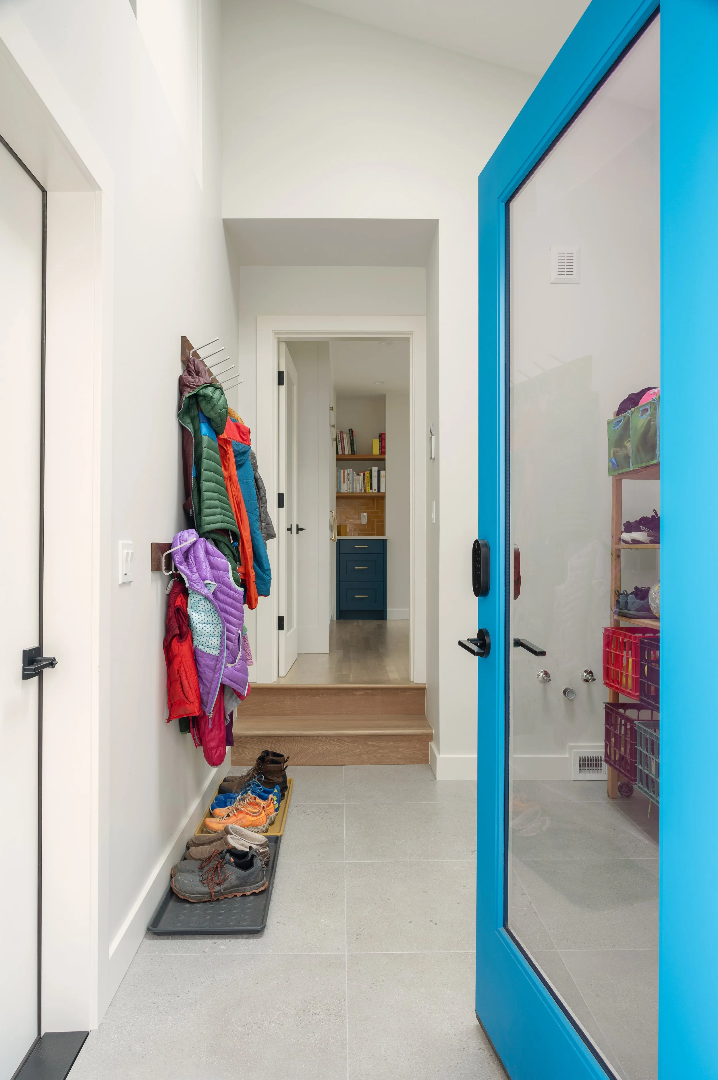 Newly remodeled mud rom entryway with colorful jackets hanging on hooks, shoes organized on a tray, and a blue door with a glass panel leading to a room with bookshelves and a countertop.