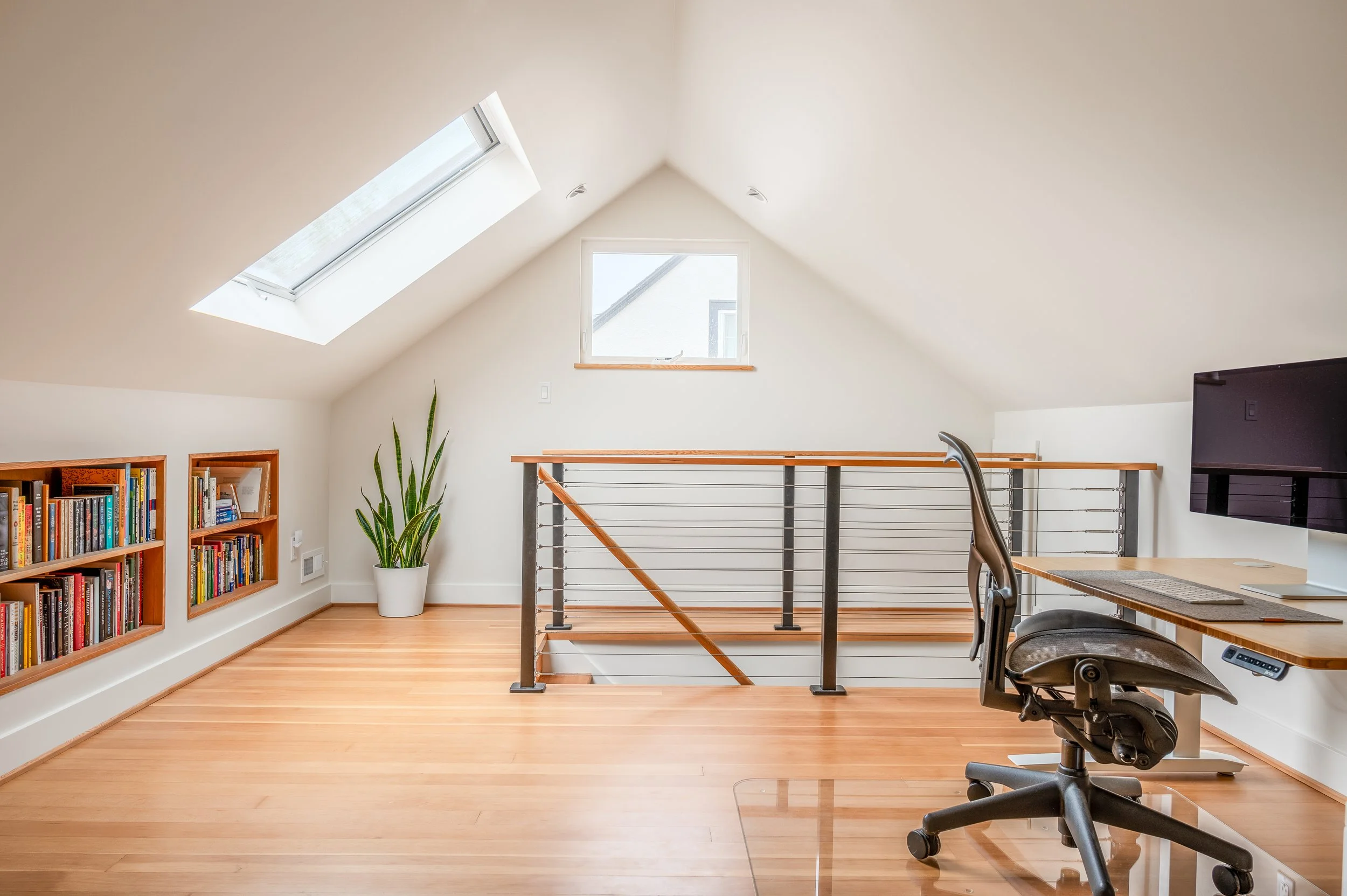 Remodeled attic room with sloped ceiling, two skylight windows, a small square window, wooden bookshelf filled with books, a potted plant, a desk with a computer, and an office chair.
