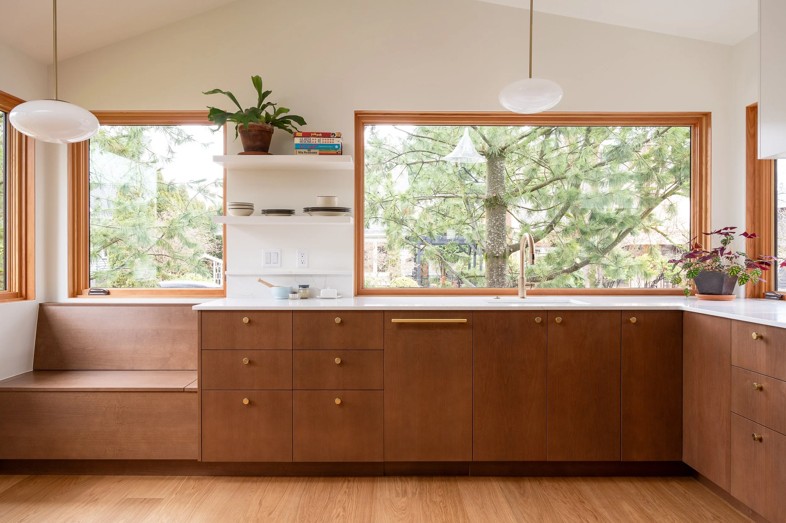 Modern, compact ADU kitchen with large windows showing trees outside, wood cabinetry, white countertops, and minimal decor including plants and stacks of dishes.