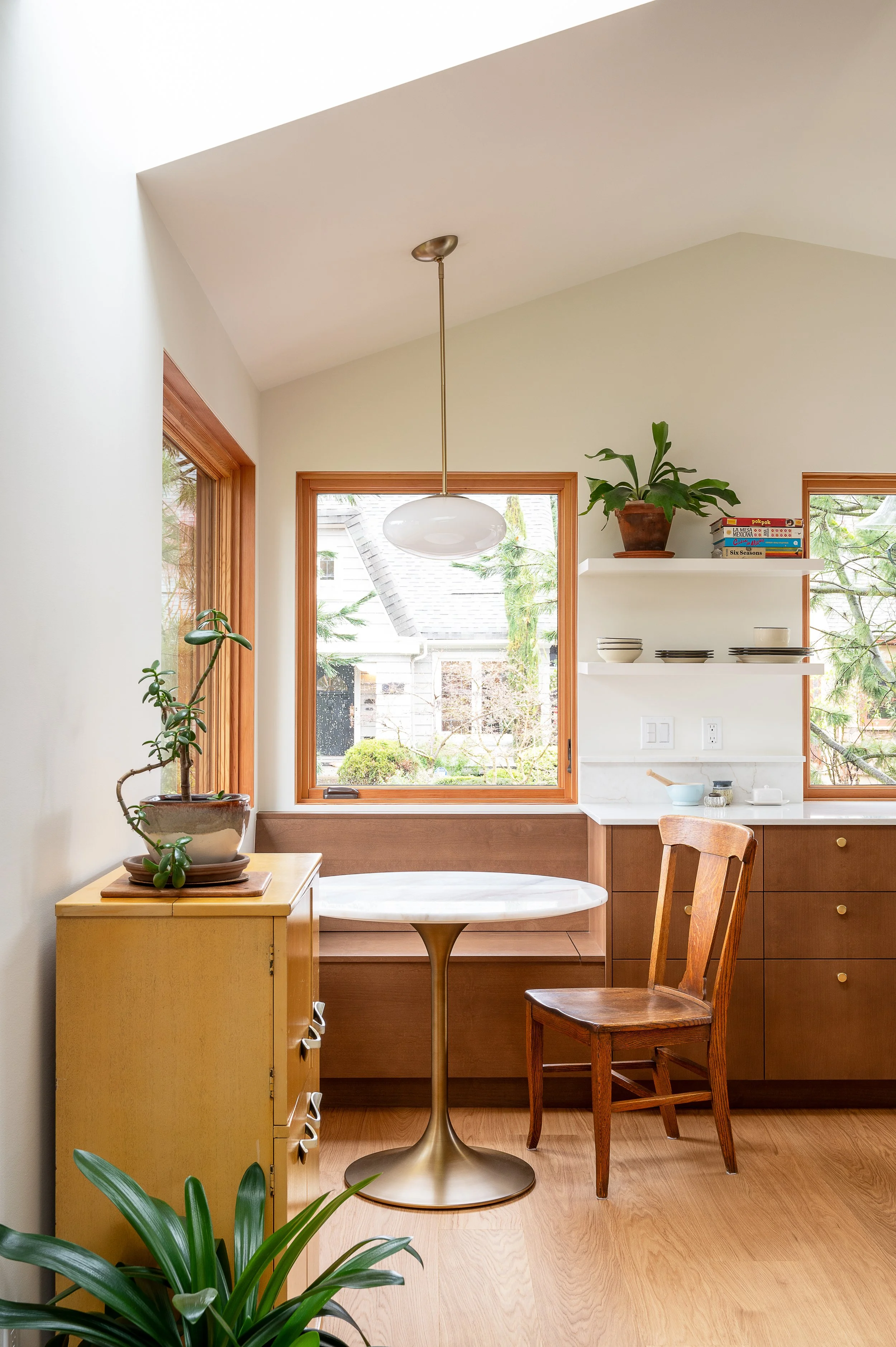 A cozy kitchen in a new ADU with large windows, a white oval dining table with a gold pedestal base, a wooden chair, a yellow cabinet, a white shelf with potted plant and books, and hardwood flooring.
