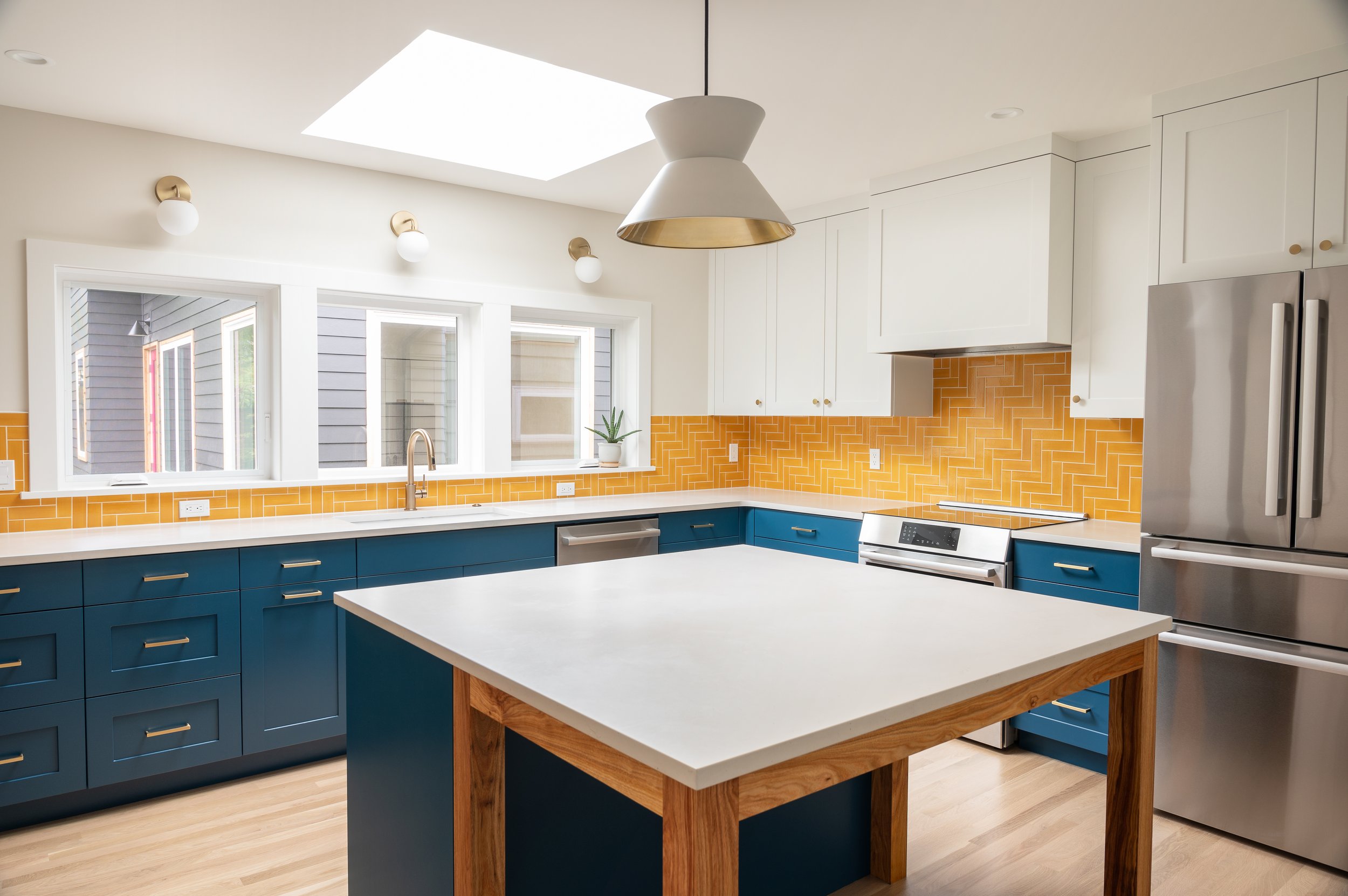 A remodeled kitchen with white cabinetry, blue lower cabinets, and a colorful yellow tile backsplash. It features a large island with a white countertop and wooden support, stainless steel appliances, and a skylight with natural light.