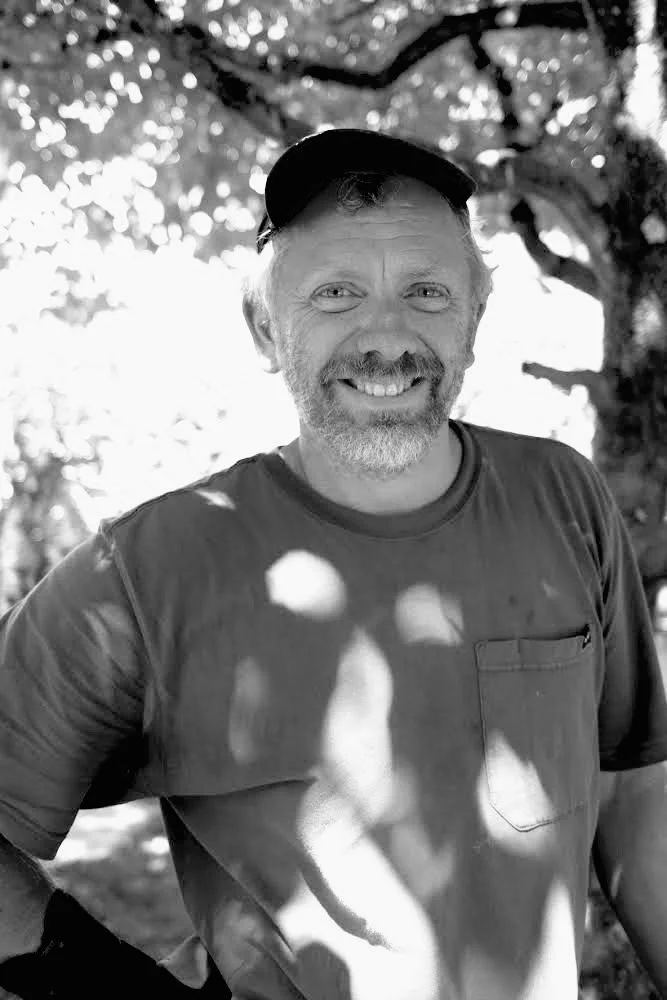 A smiling man with a beard and mustache, wearing a baseball cap and a T-shirt, standing outdoors with trees in the background.