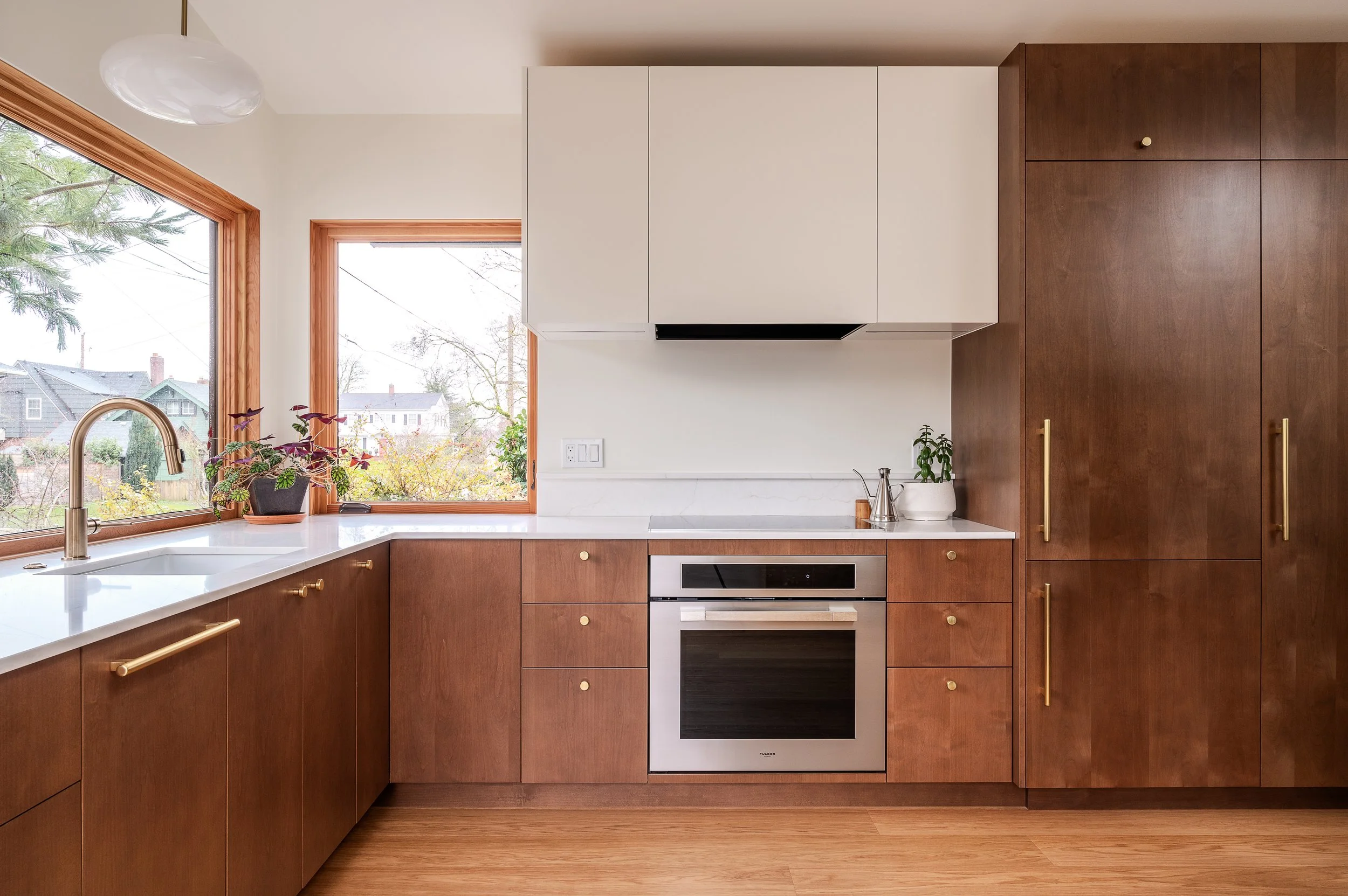ADU Kitchen with wooden cabinets, white countertops, a window with a view of trees and houses, a potted plant on the windowsill, and a built-in oven.