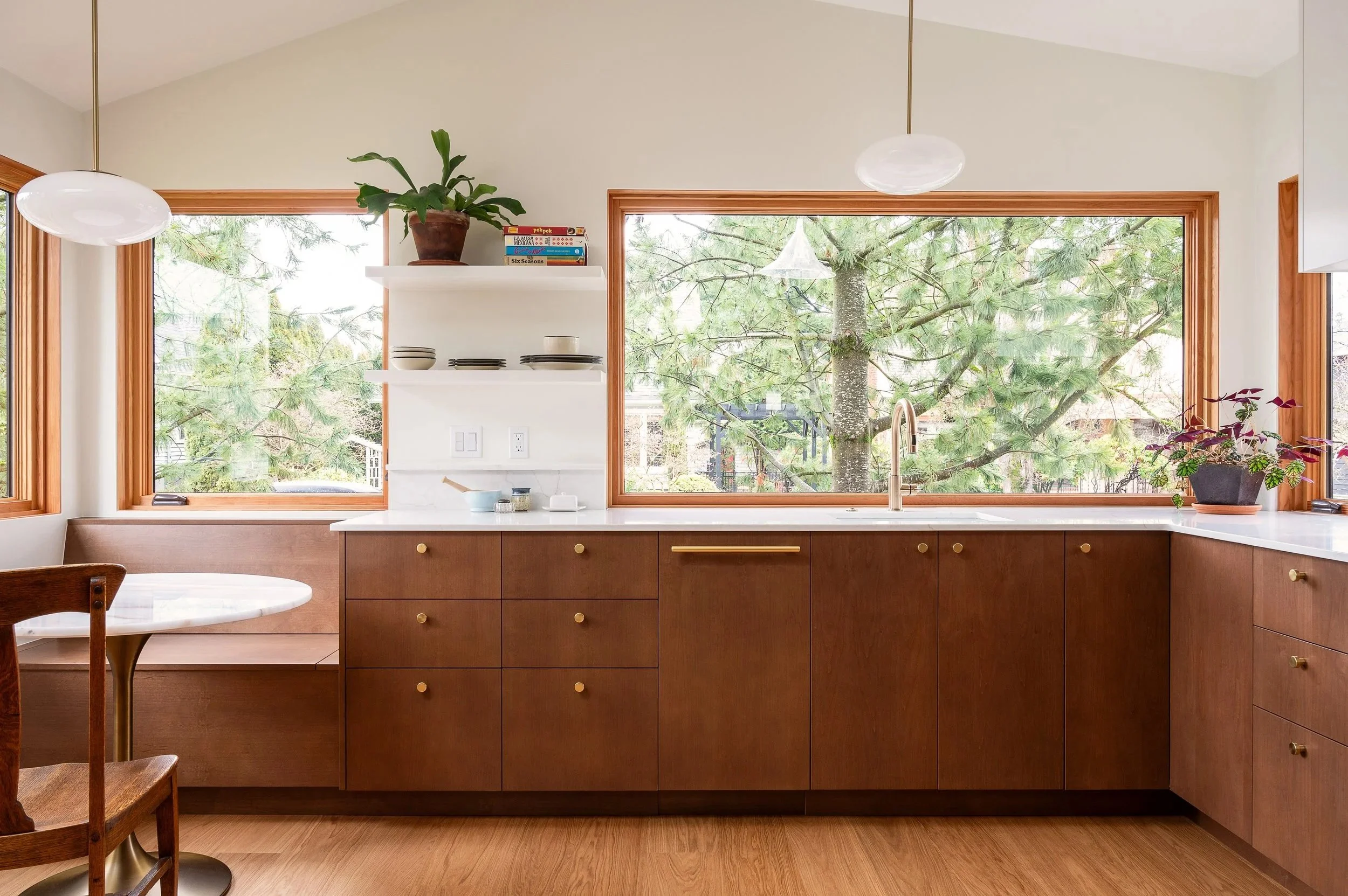 Kitchen with wooden cabinets, large windows showing a tree outside, white countertops, and minimal decor.