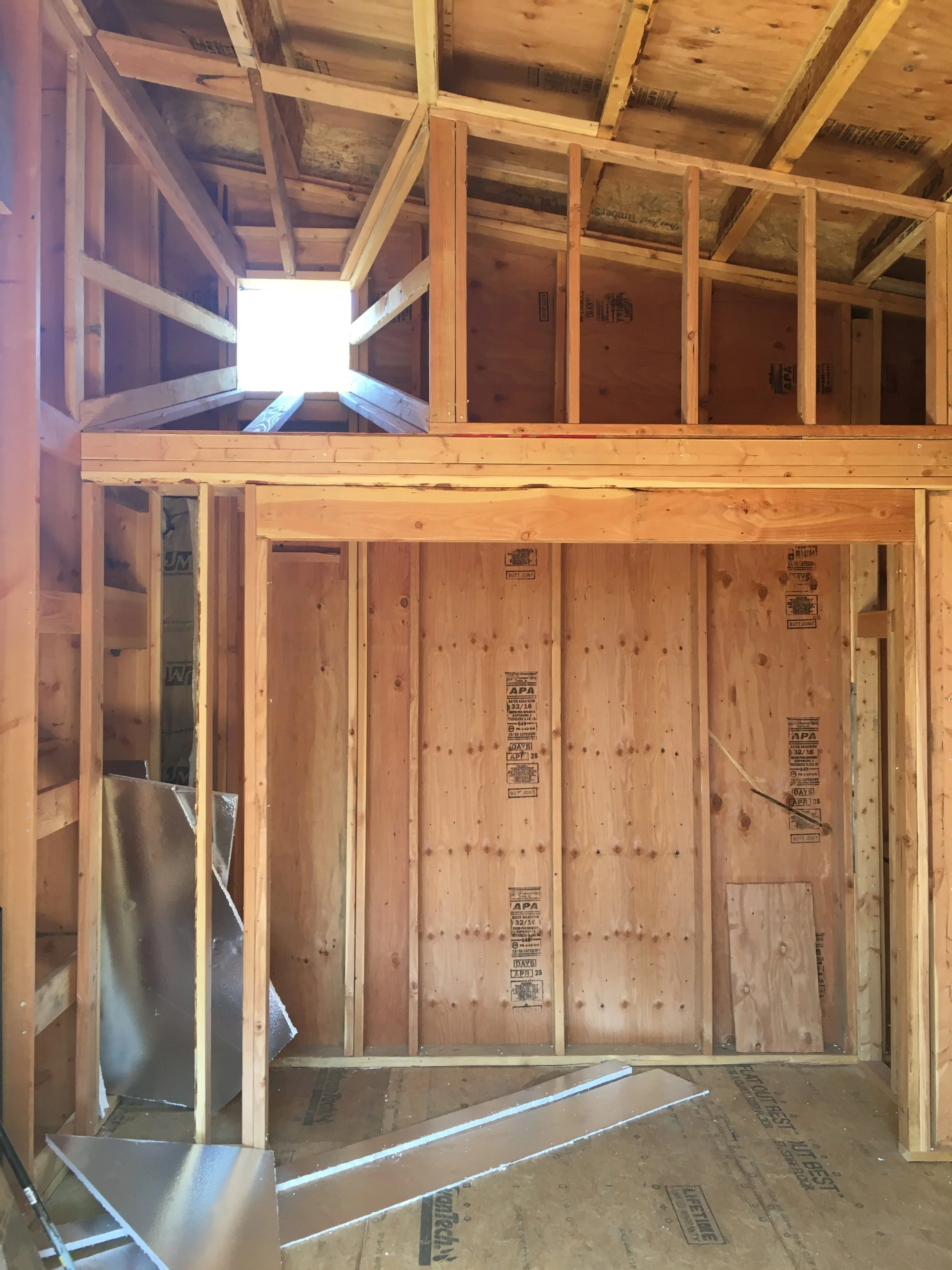 Interior view of a building under construction with wooden framing, partially installed wall panels, and a skylight opening at the top.