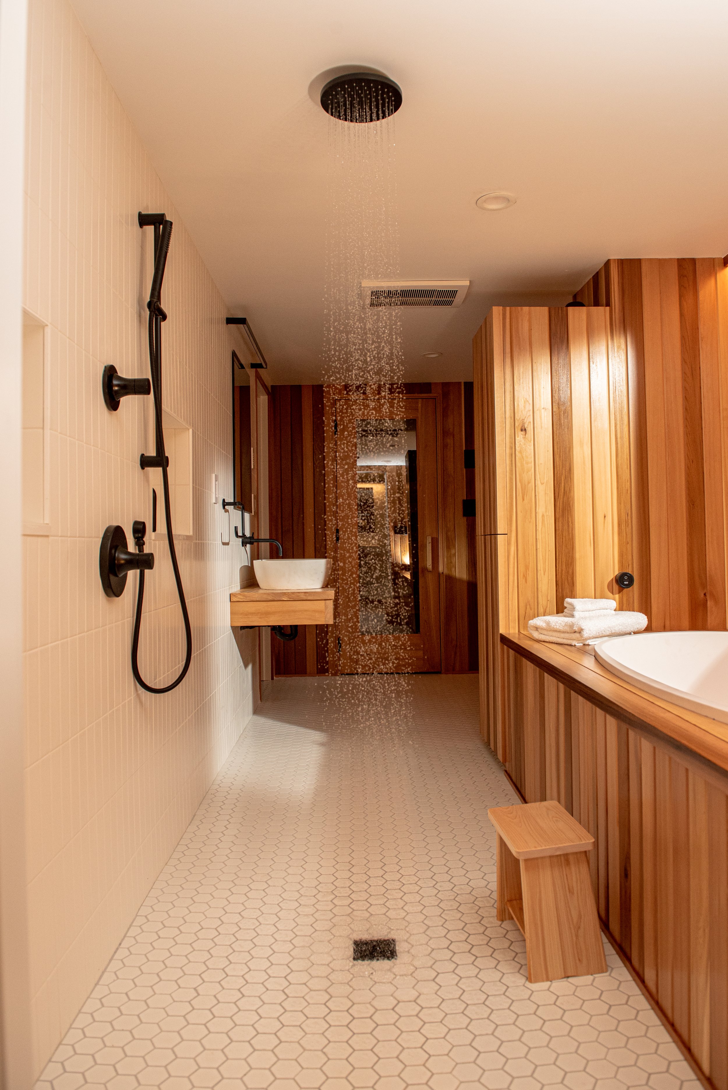 Bathroom with wooden paneling, a shower with a rain showerhead, a vessel sink on a wooden floating vanity, neatly folded towels near a bathtub, hexagonal floor tiles, and a small wooden stool.