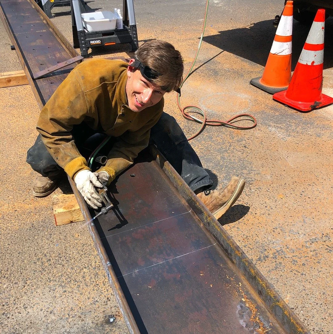 A construction worker is kneeling on the ground, welding a metal beam with safety goggles on. Bright orange traffic cones and construction equipment are visible in the background.