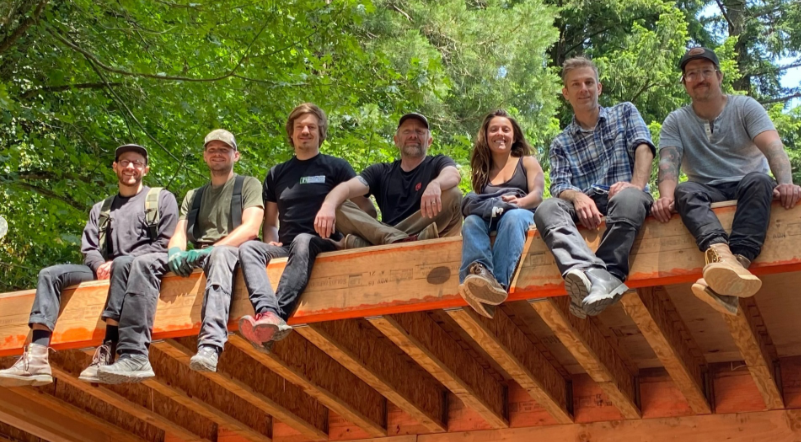 Group of seven people sitting on a wooden structure under green leafy trees outdoors.