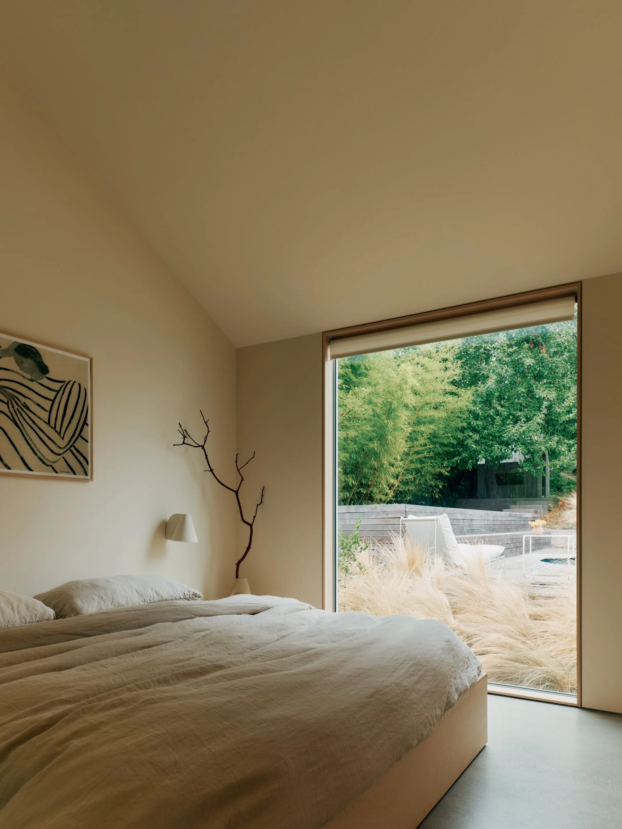 A bedroom in a pool house with rental space with a large bed covered in cream-colored linens, a split window showing greenery outside, a framed artwork on the wall, a small bedside lamp, and a decorative branch inside the room.