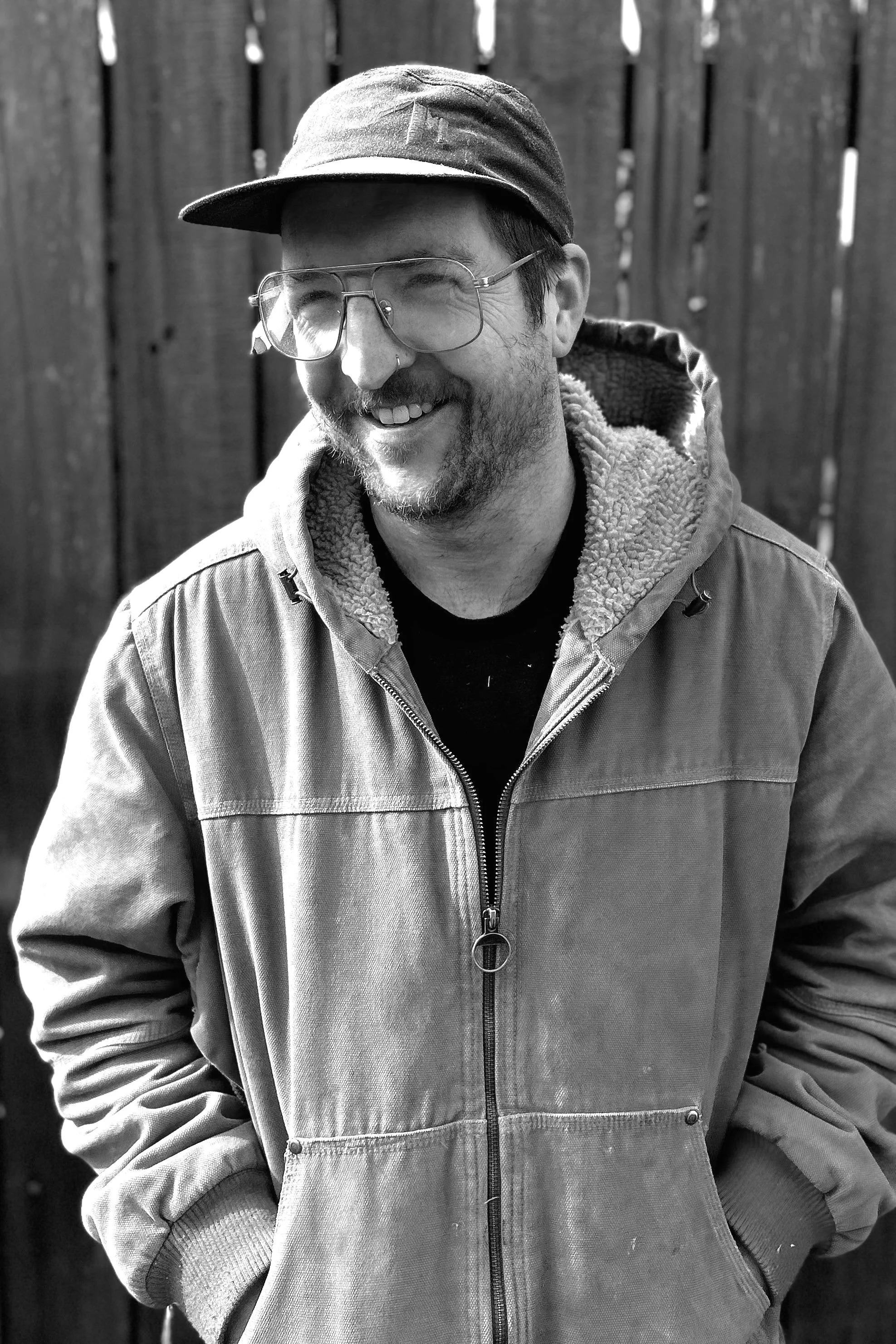 A man wearing glasses, a cap, and a hooded jacket, smiling, in front of a wooden fence.