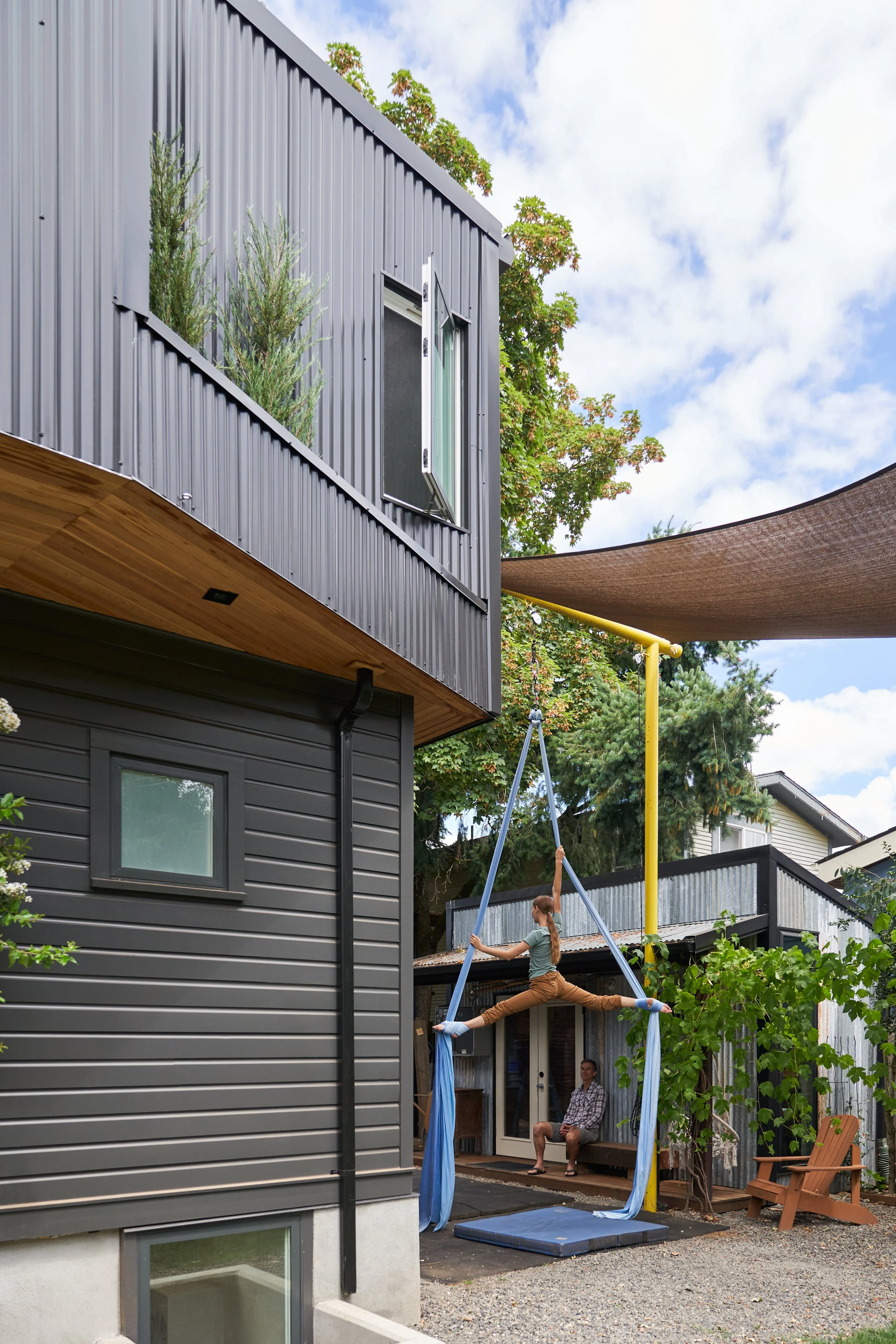 A woman performs a split on a trapeze in a backyard with a man seated nearby, surrounded by modern houses and trees.