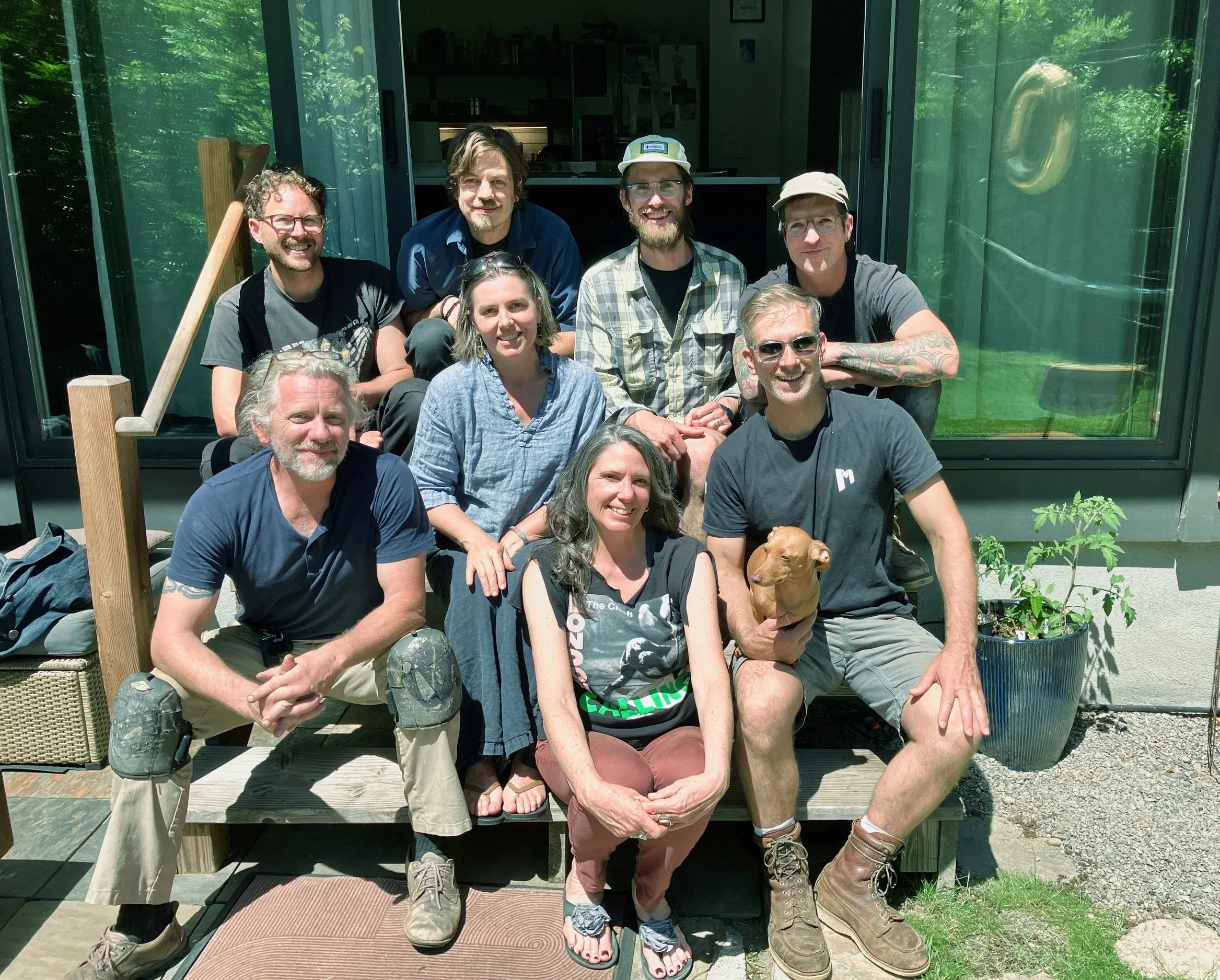 A group of nine people, seven men and two women, posed together outdoors on a wooden deck in front of a green glass building with reflections of trees. They are smiling and sitting or standing closely. One dog is lying at the front.