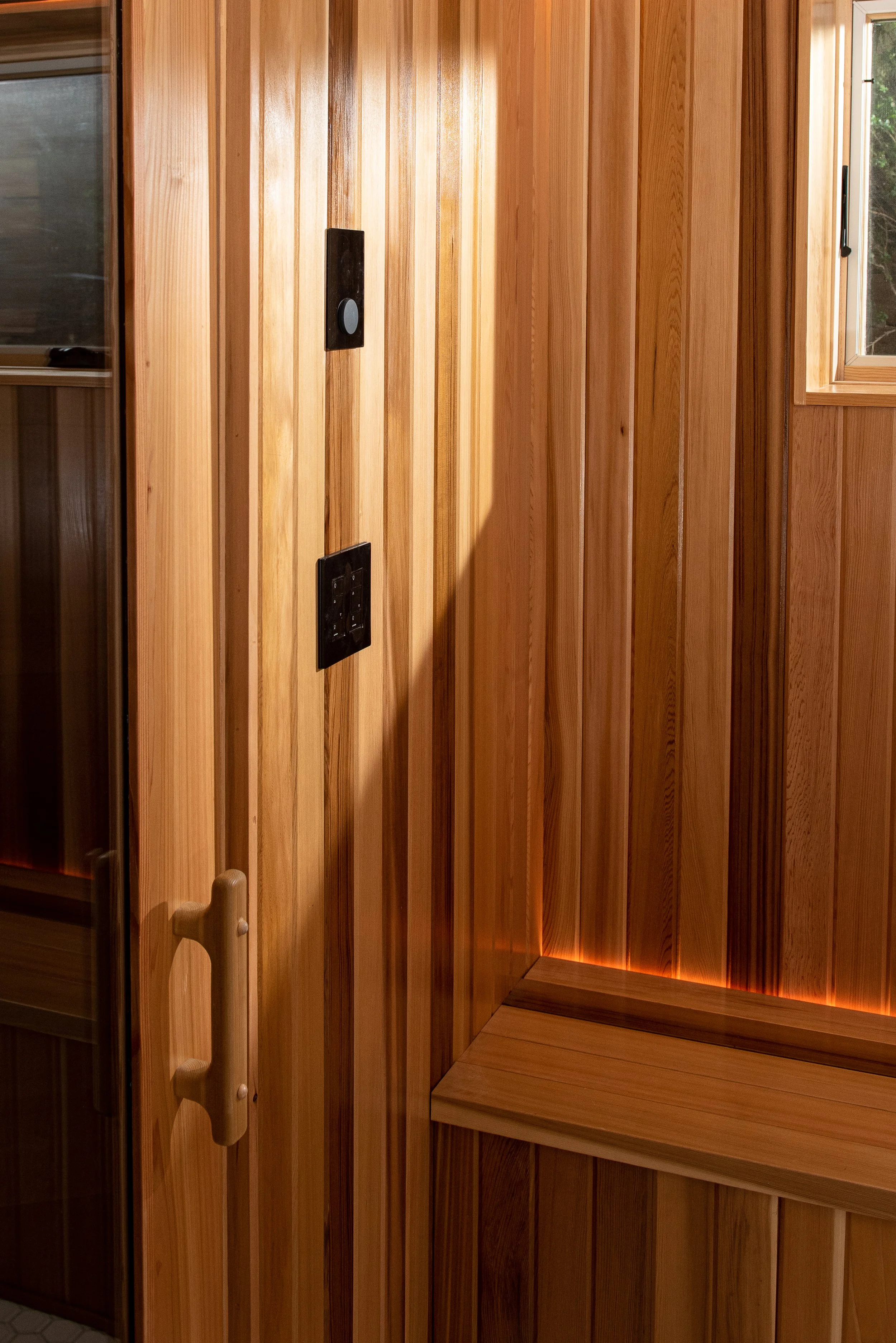 Outside the glass door of the cedar sauna basement renovation is a cedar bench, with rectangular widow above, and unobtrusive yet helpful light coming from the crack where bench meets wall.