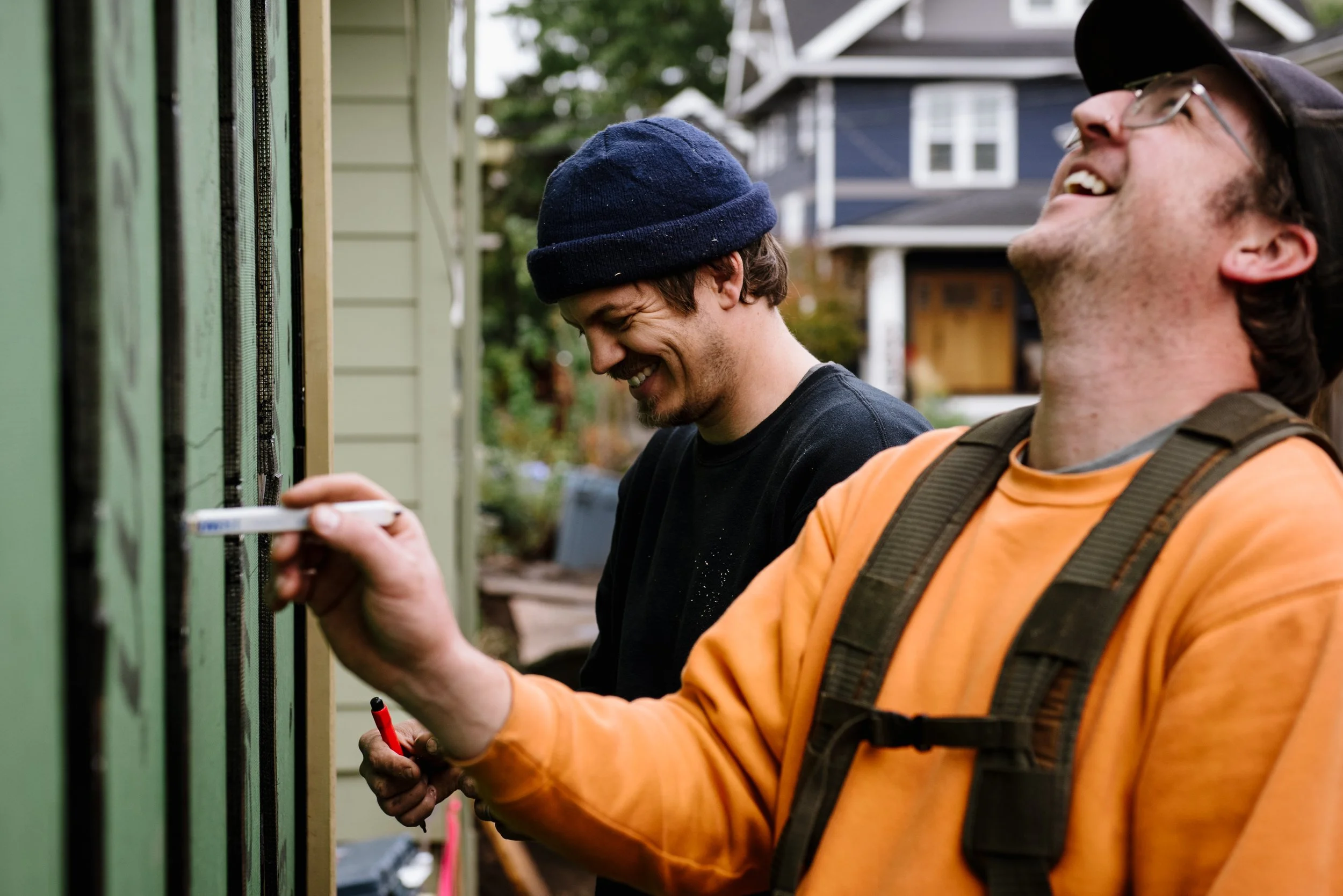 Two men laughing and working on an outdoor electrical box. One man is wearing an orange shirt and a hat, and is holding a screwdriver. The other man is wearing a black shirt and a beanie.