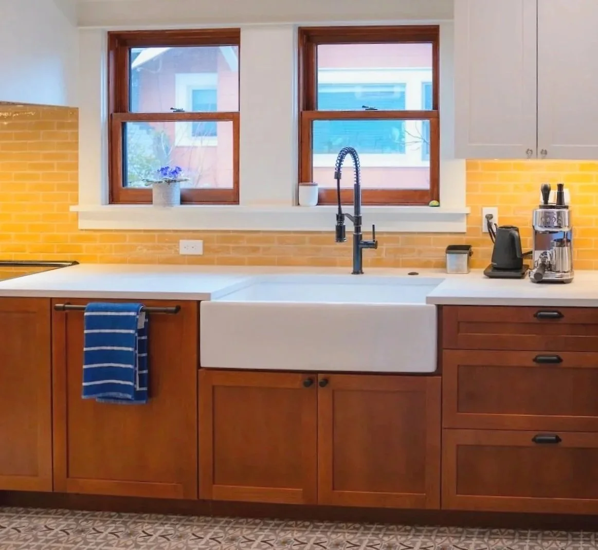 On the left is a stainless steel oven, white countertop, rectangular sink basin built into the dark wood cabinets, drawers and dark metal hardware and fixtures.