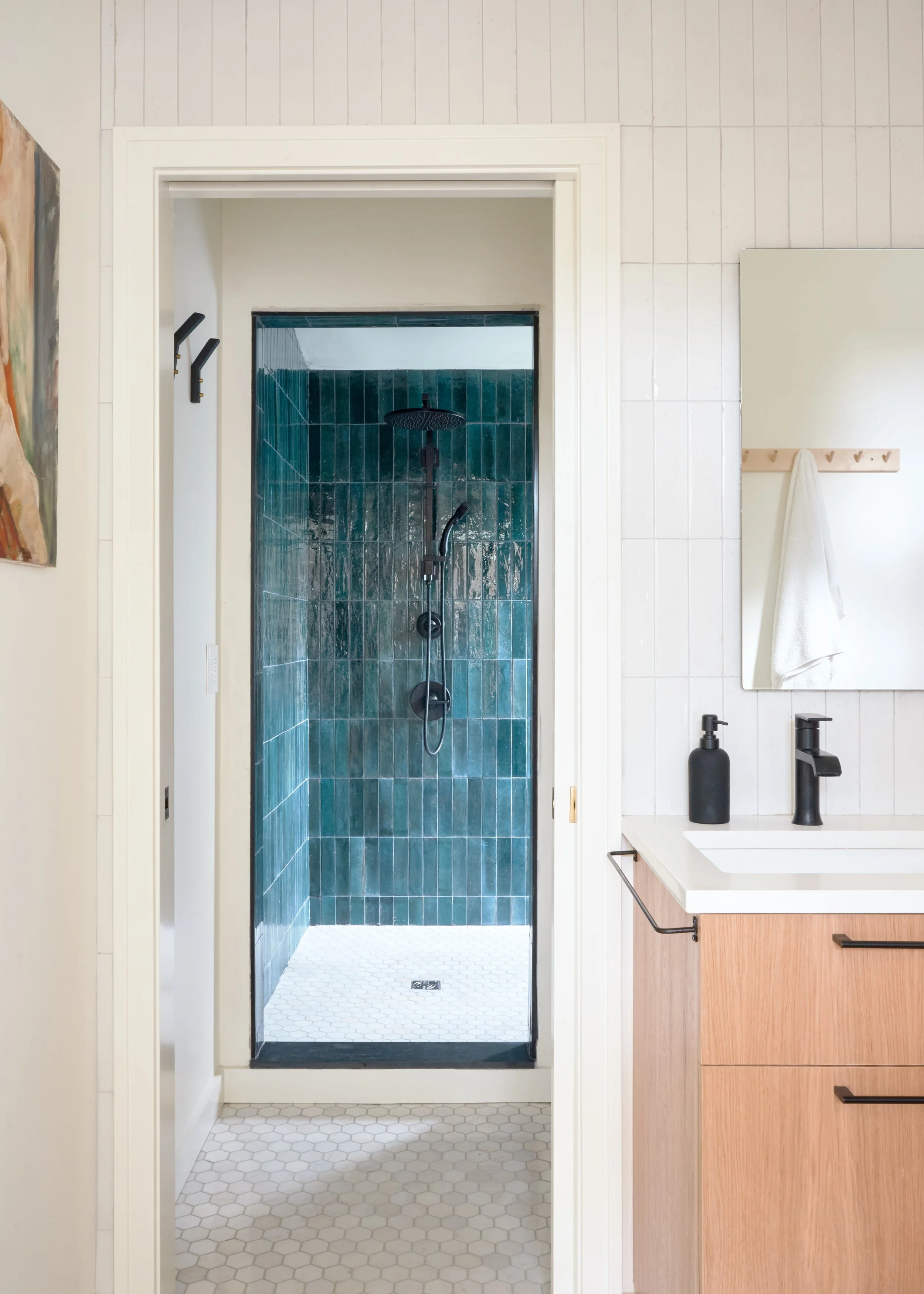 Indoor bathroom showing a view into a shower with green tiles, a black showerhead, and white hexagonal floor tiles, next to a vanity with a black soap dispenser and faucet, and a mirror with a white towel hanging.