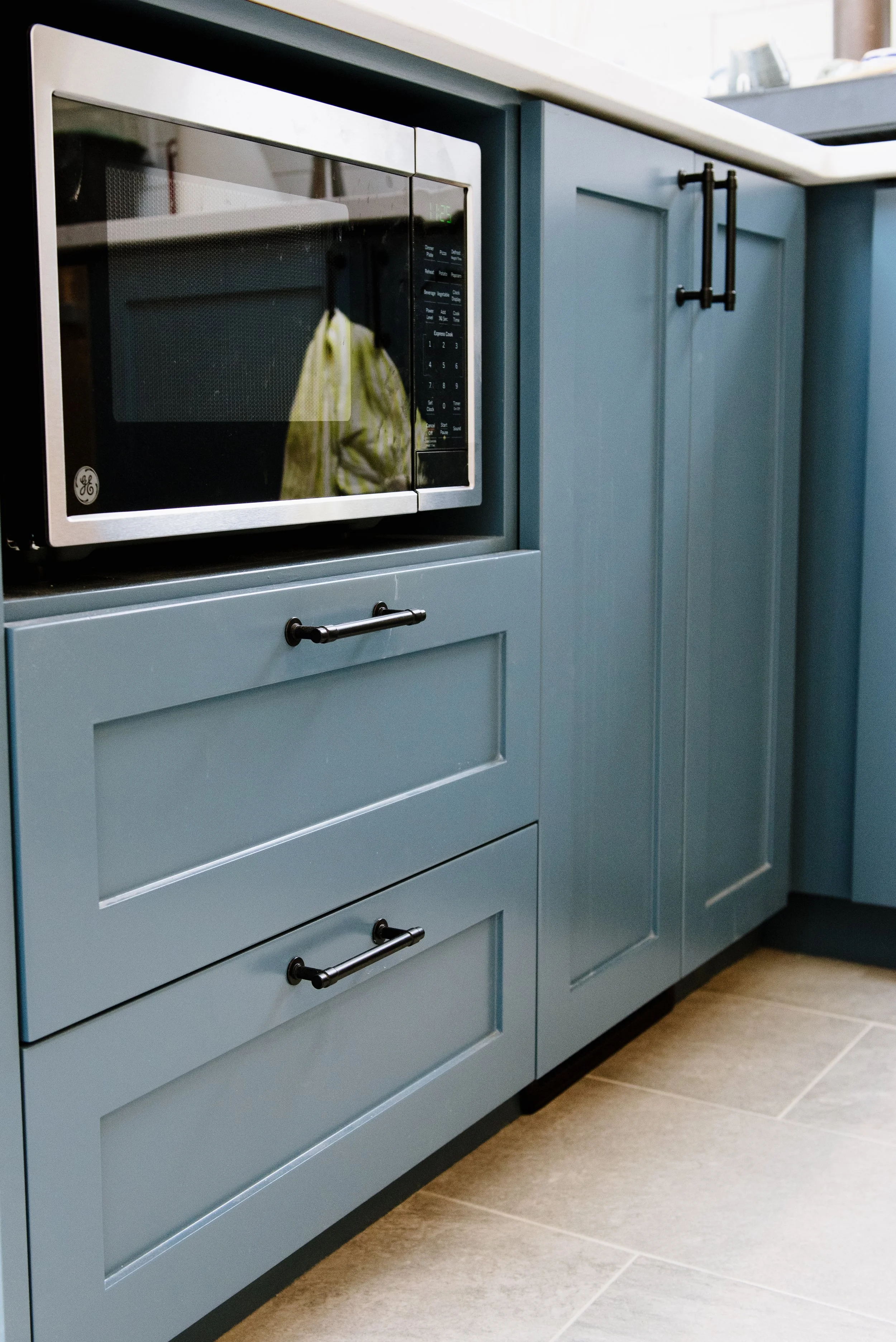 Close-up of a microwave oven built into remodeled blue kitchen cabinetry with black hardware and beige tile flooring.