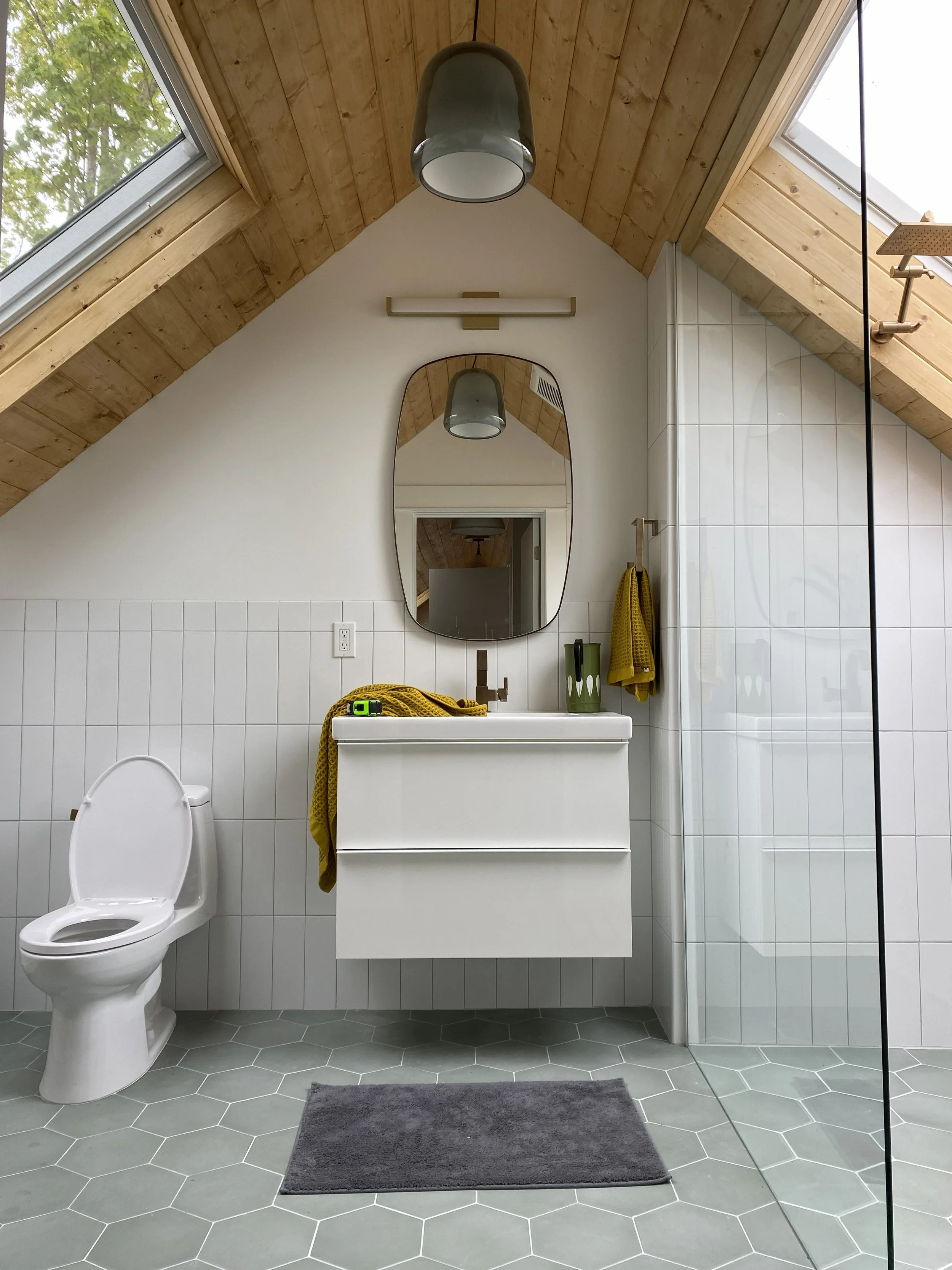 2 skylights add natural light to this small attic remodel bathroom, one over the toilet on the left, one over the shower on the right with sink in the middle. Walls have rectangle white tile and with rounded cornered rectangle mirror above the sink.