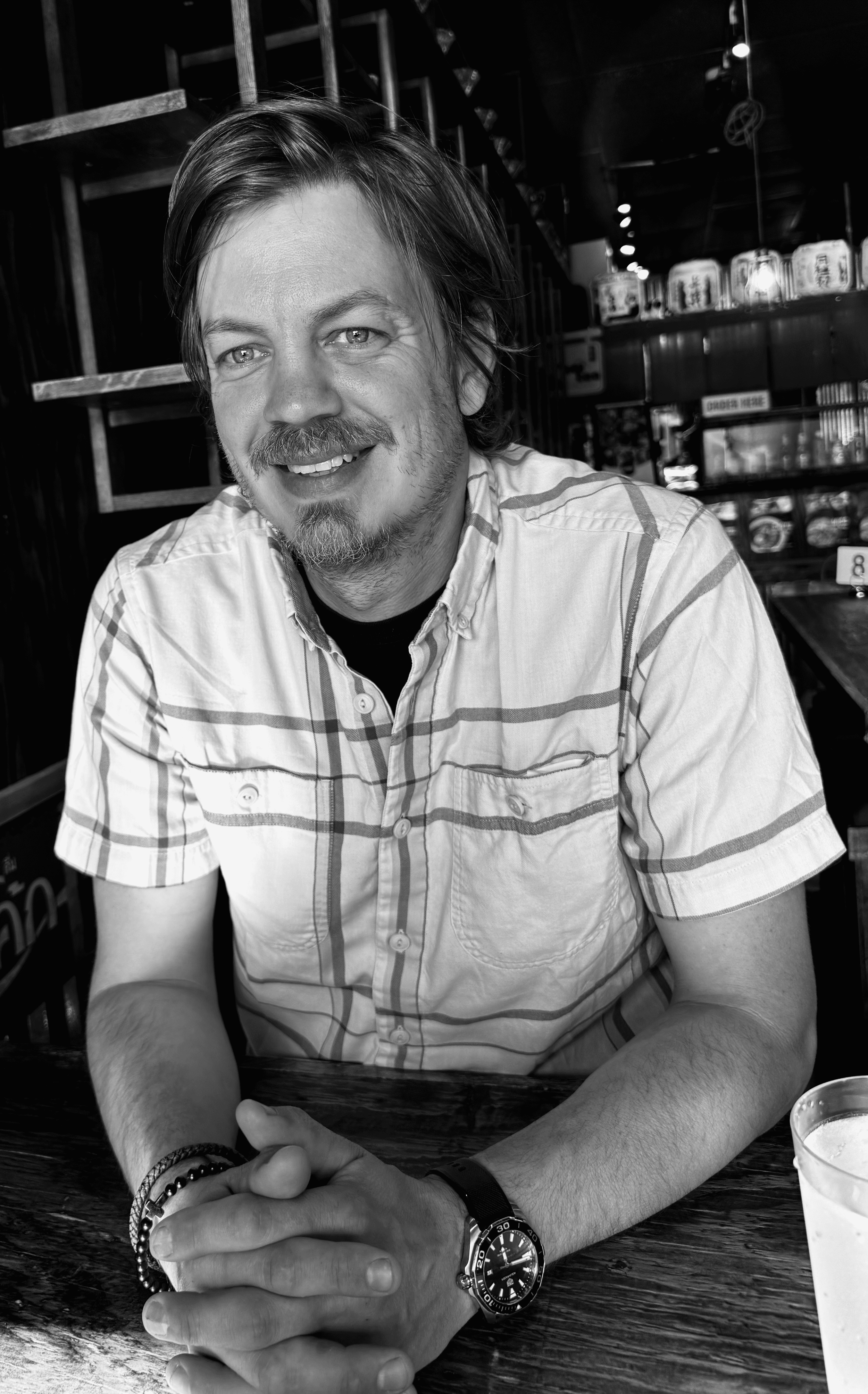 A man with light skin, short tousled hair, a mustache and goatee, smiling with crossed arms, wearing a black long-sleeve shirt and a wristwatch, standing in front of a wooden fence.