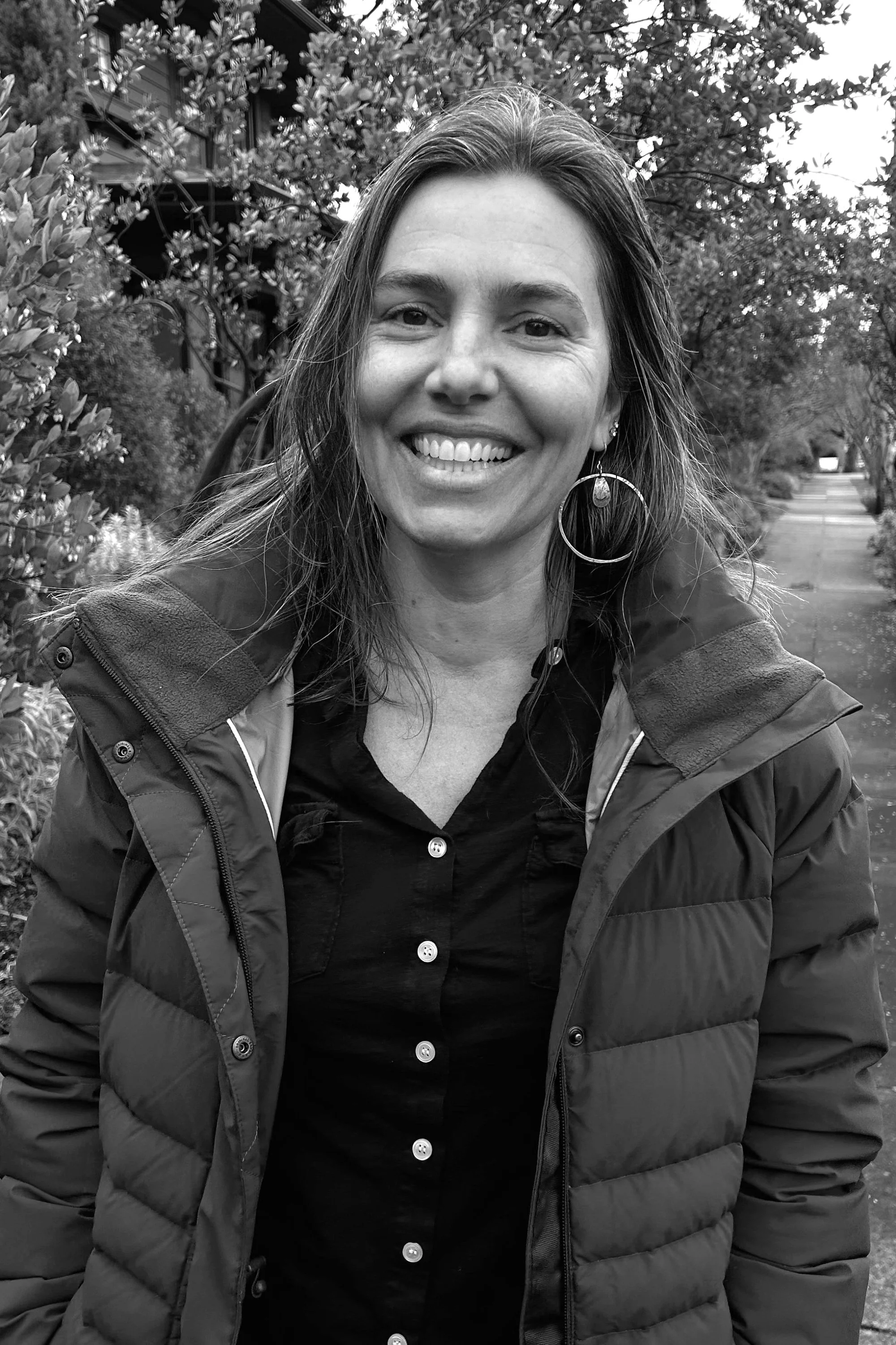 A woman smiling outdoors in a black and white photo, wearing a jacket and hoop earrings, standing on a sidewalk with trees and houses in the background.