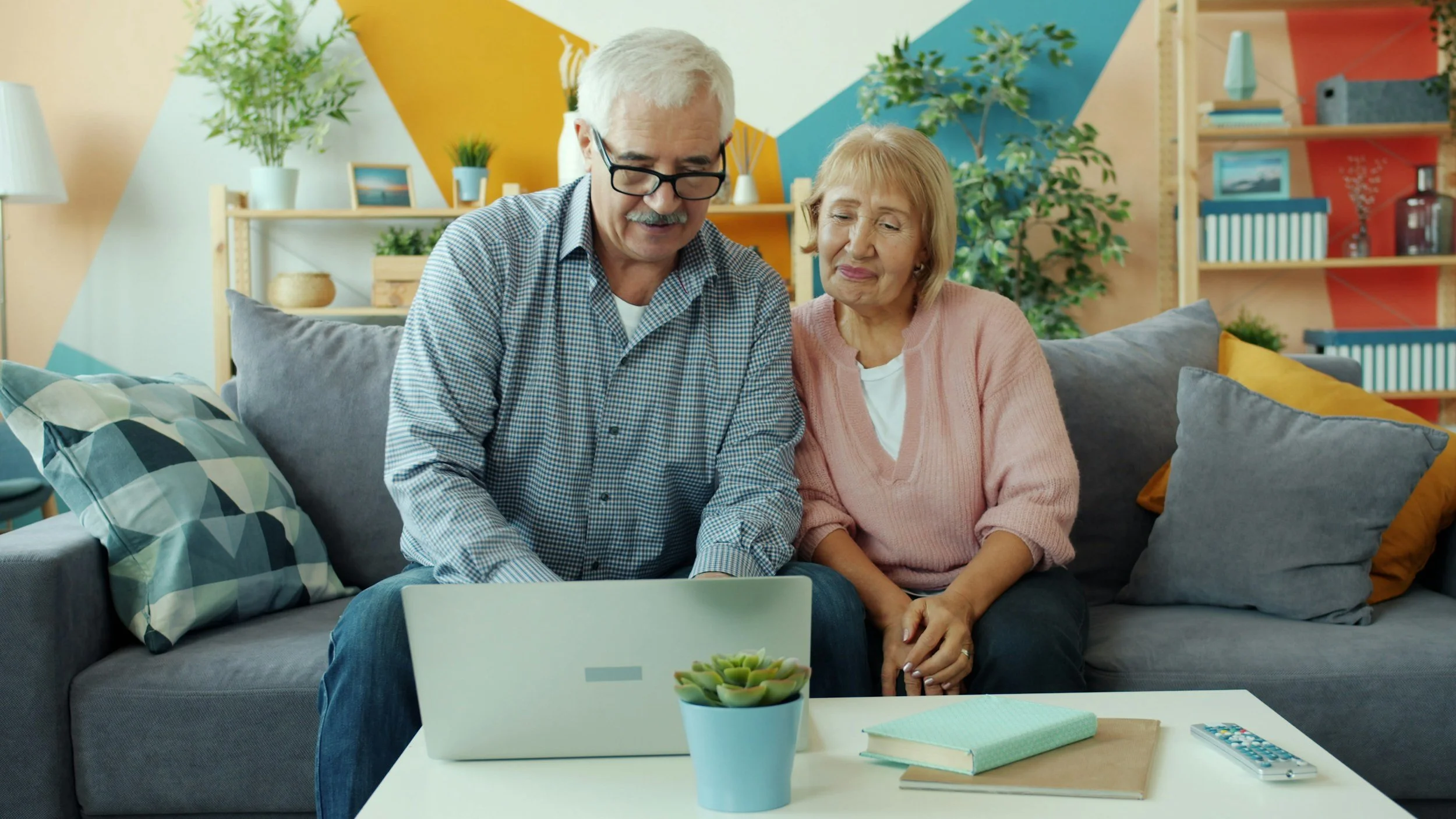A man with white hair and glasses sitting on a couch with a woman with blond hair. They are looking at a laptop.