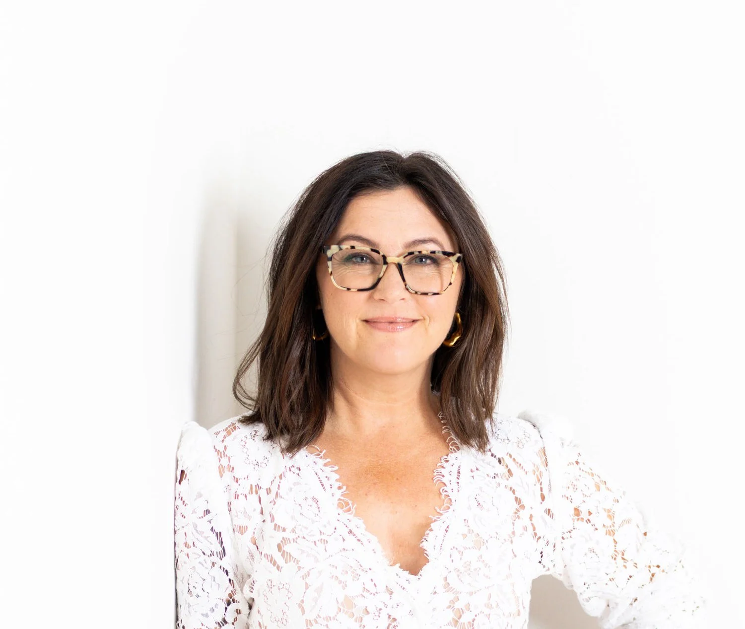 A woman with shoulder-length brown hair, glasses, and gold earrings, smiling while wearing a white lace top, standing against a plain white background.